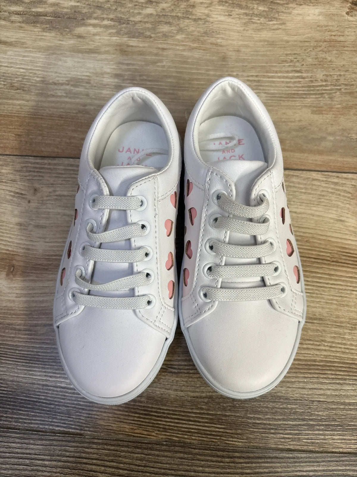 Top view of Janie & Jack heart sneakers showcasing white uppers, pink heart details, and bungee laces on wood grain