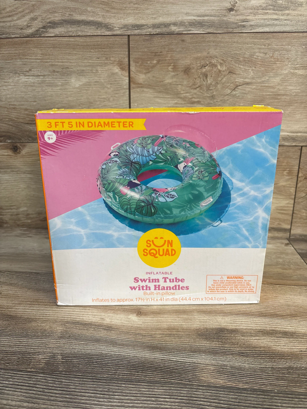 Front view of boxed Sun Squad swim tube with handles showing tropical leaf pattern and 3 ft 5 in diameter label on wood floor