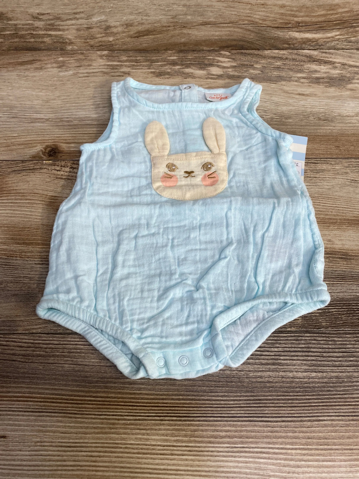 Front view of Cat and Jack light blue gauze baby romper showcasing embroidered bunny applique and three snap crotch closures on wood background