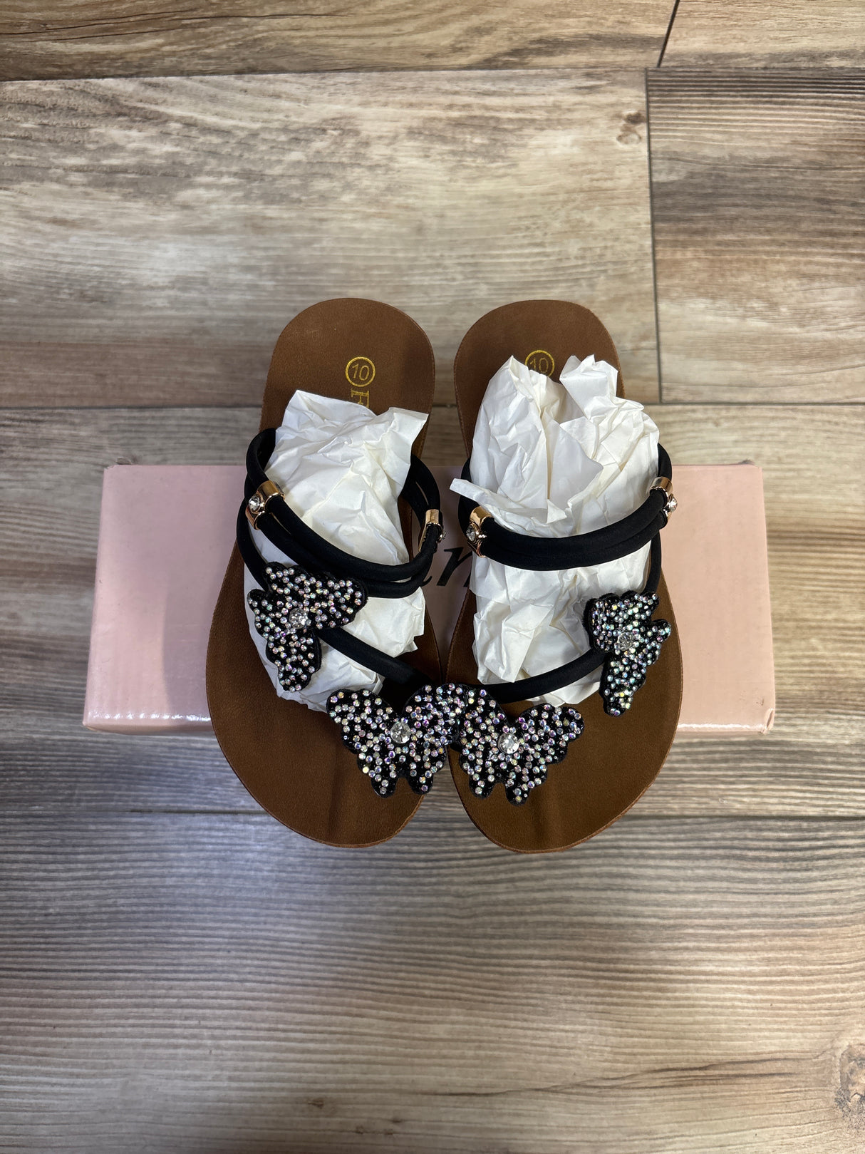 Top-down view of brown sandals with black straps and 3 rhinestone butterfly decorations, size 10C visible on a pink box