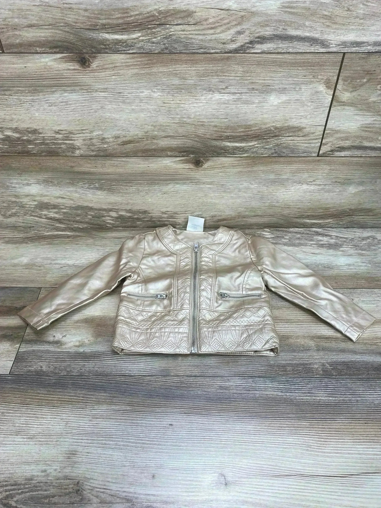 Flat lay of toddler Genuine Kids champagne faux leather jacket showing front zipper, quilted stitched panels and zip pockets on wood floor
