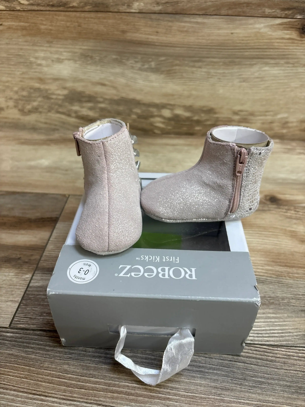 Three-quarter view of Robeez First Kicks Madison baby boots showcasing shimmery pink glitter, side zipper, and textured heel on a gray Robeez 0-3 months box on wood plank floor