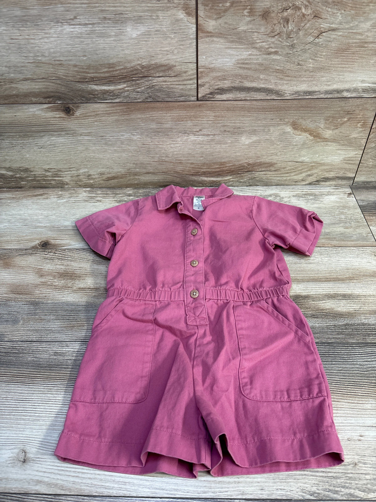 Flat lay of Oshkosh pink polo shortie romper size 3T showing front buttons, elastic waist, and pockets on wood background