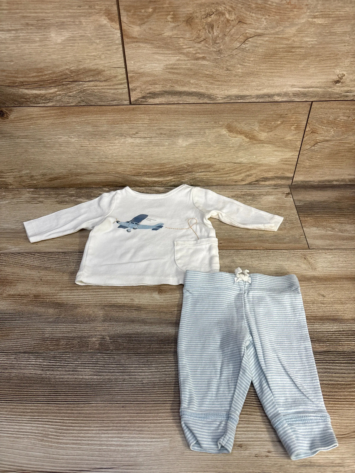 Front view of Carter’s two-piece shirt and pants set showcasing an airplane graphic, pocket, and striped pants on a wood floor
