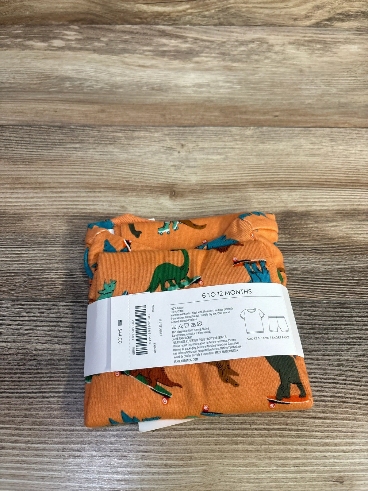 Top-down of Janie and Jack Dino Skate 2pc orange pajamas with dinosaur-on-skateboard print and size label 6 TO 12 MONTHS on wood