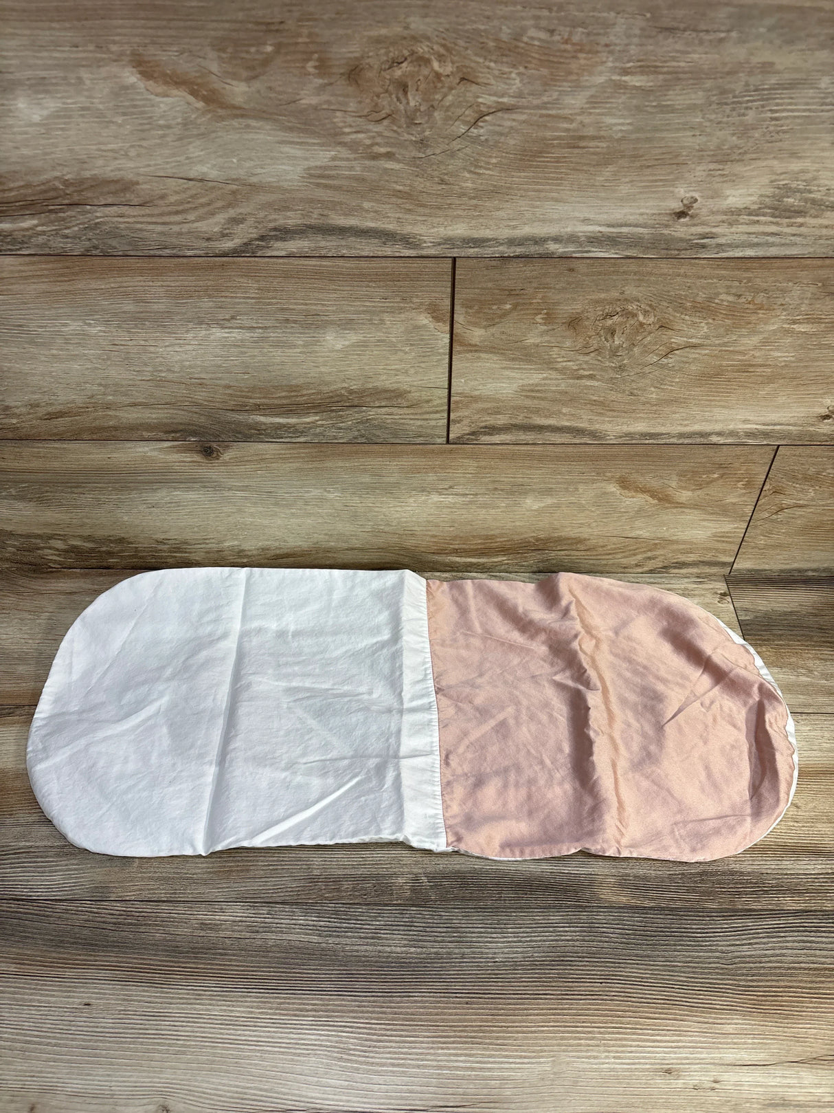 Flat lay of Silky Tots Silk oval bassinet sheet in pink showing two-tone white and light pink panels on wooden floor