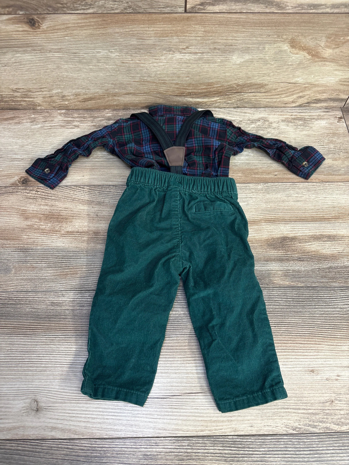Toddler outfit with green corduroy pants, plaid shirt, and suspenders on wood floor