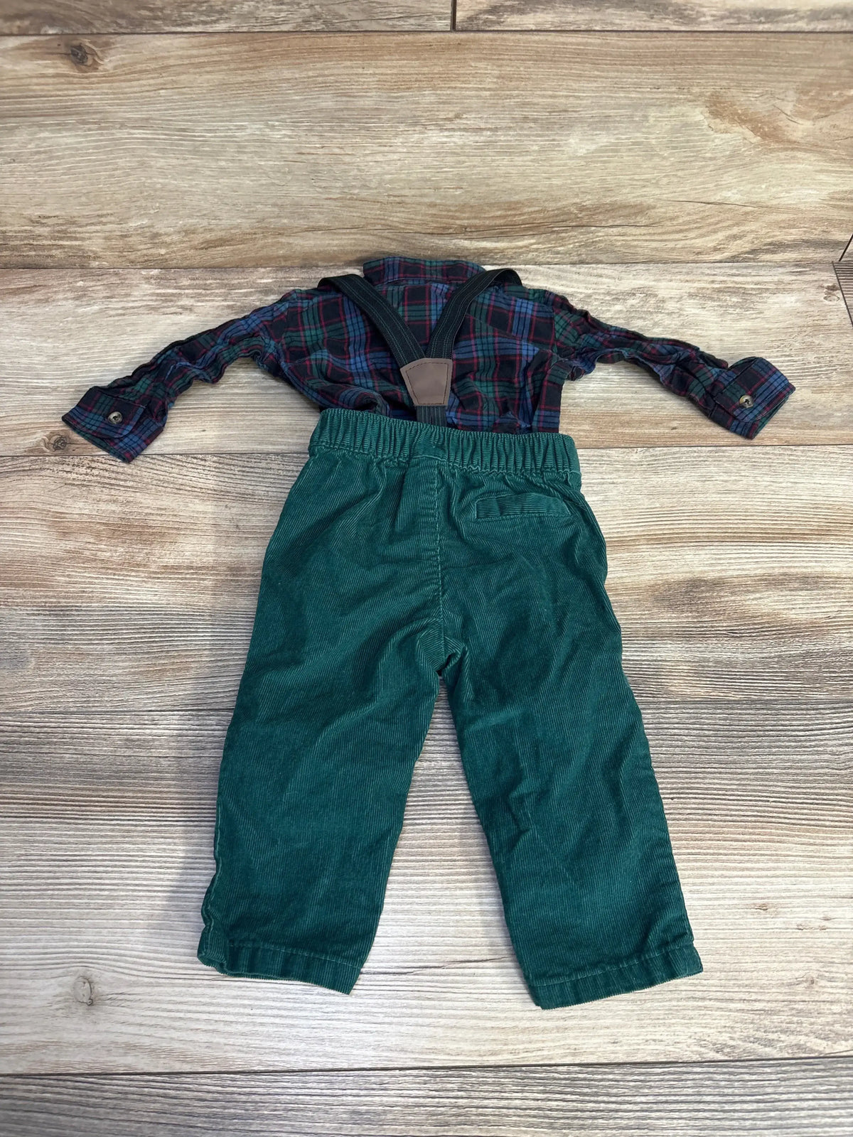 Toddler outfit with green corduroy pants, plaid shirt, and suspenders on wood floor