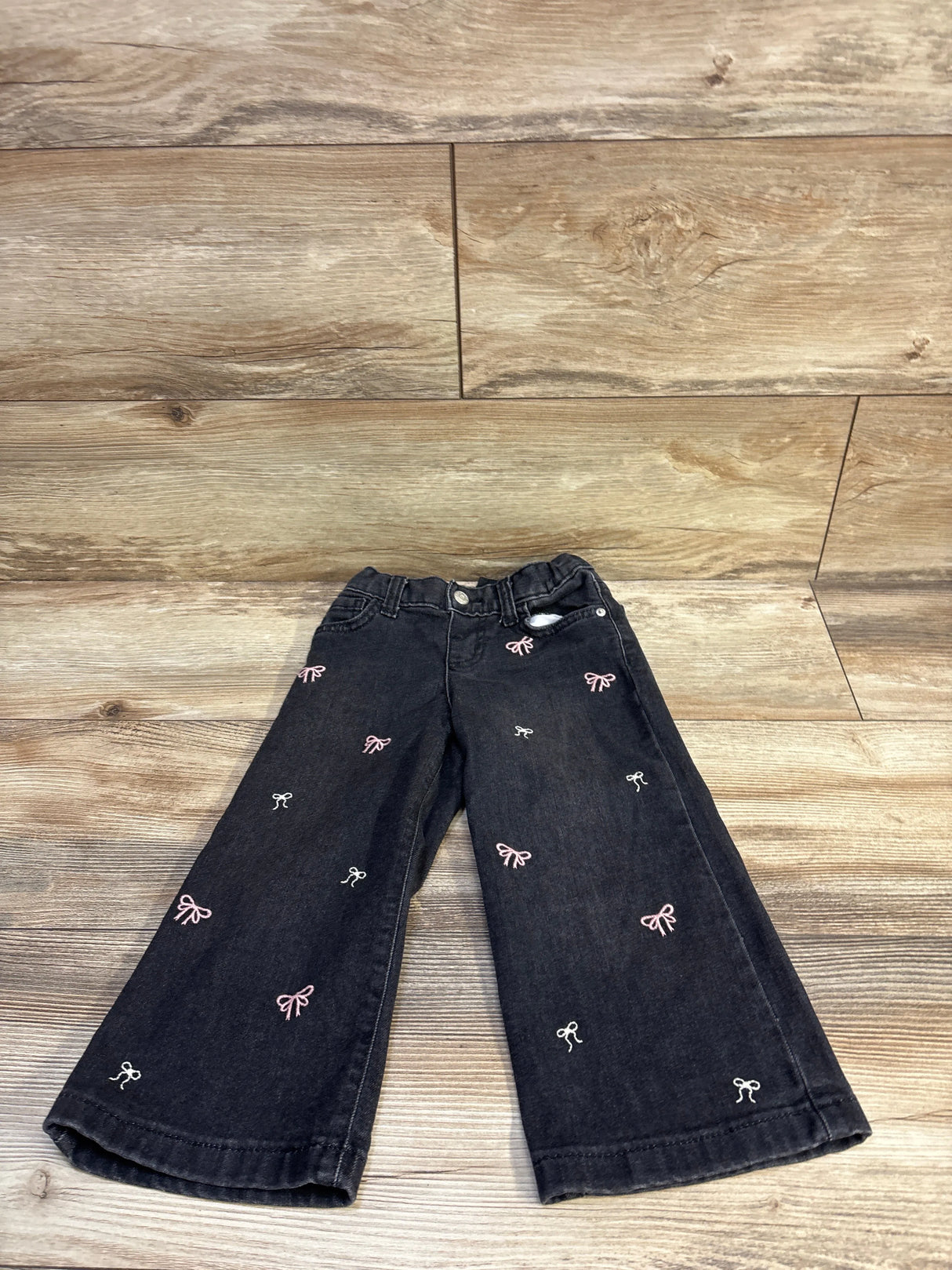 Front flat lay of toddler black wide-leg jeans showcasing scattered pink and white embroidered bows with light wood background