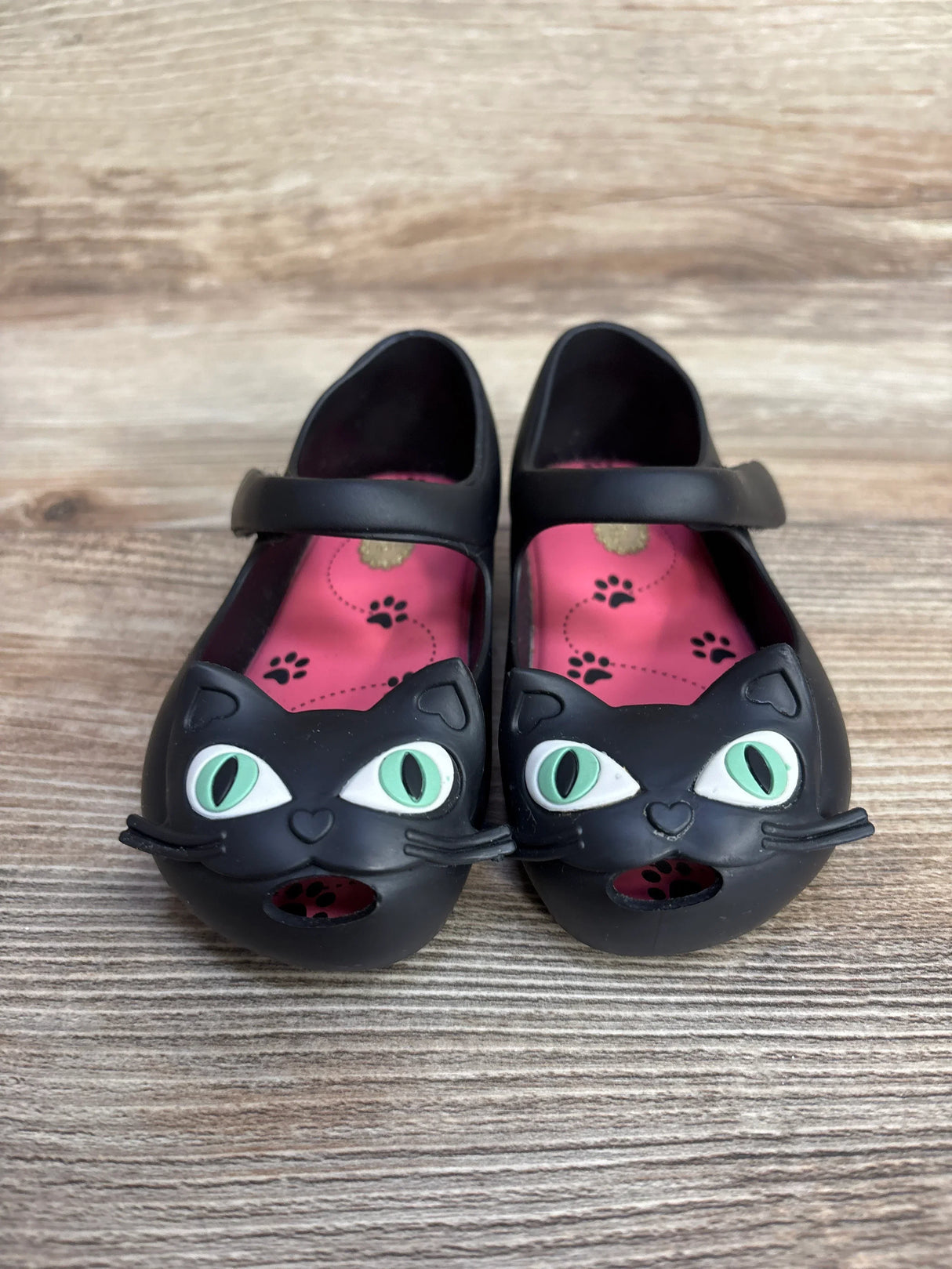 Pair of black children's shoes with green-eyed cat face design and pink paw print insoles on wooden floor