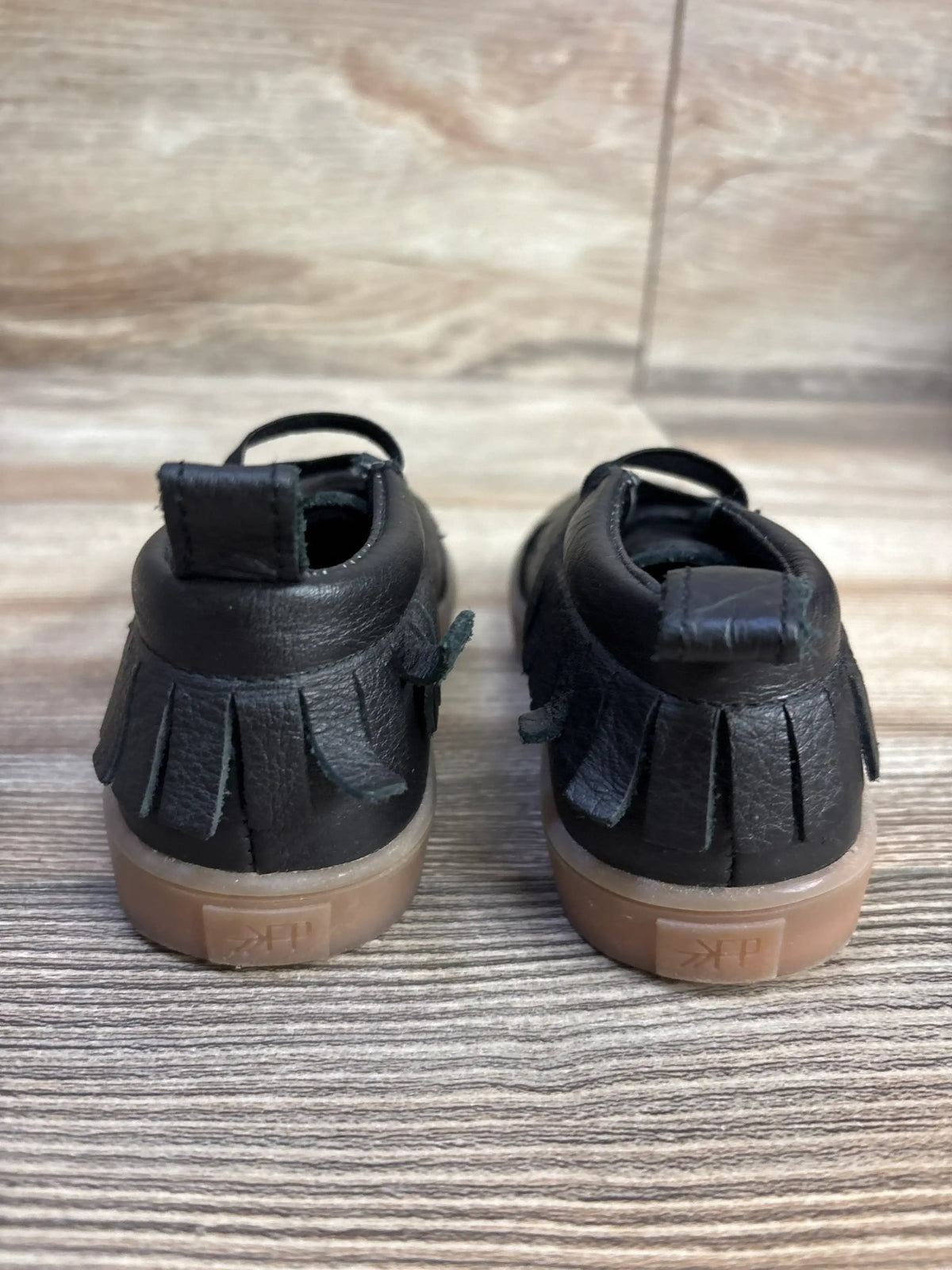 Black leather baby moccasins with fringe, gum soles, on wood background