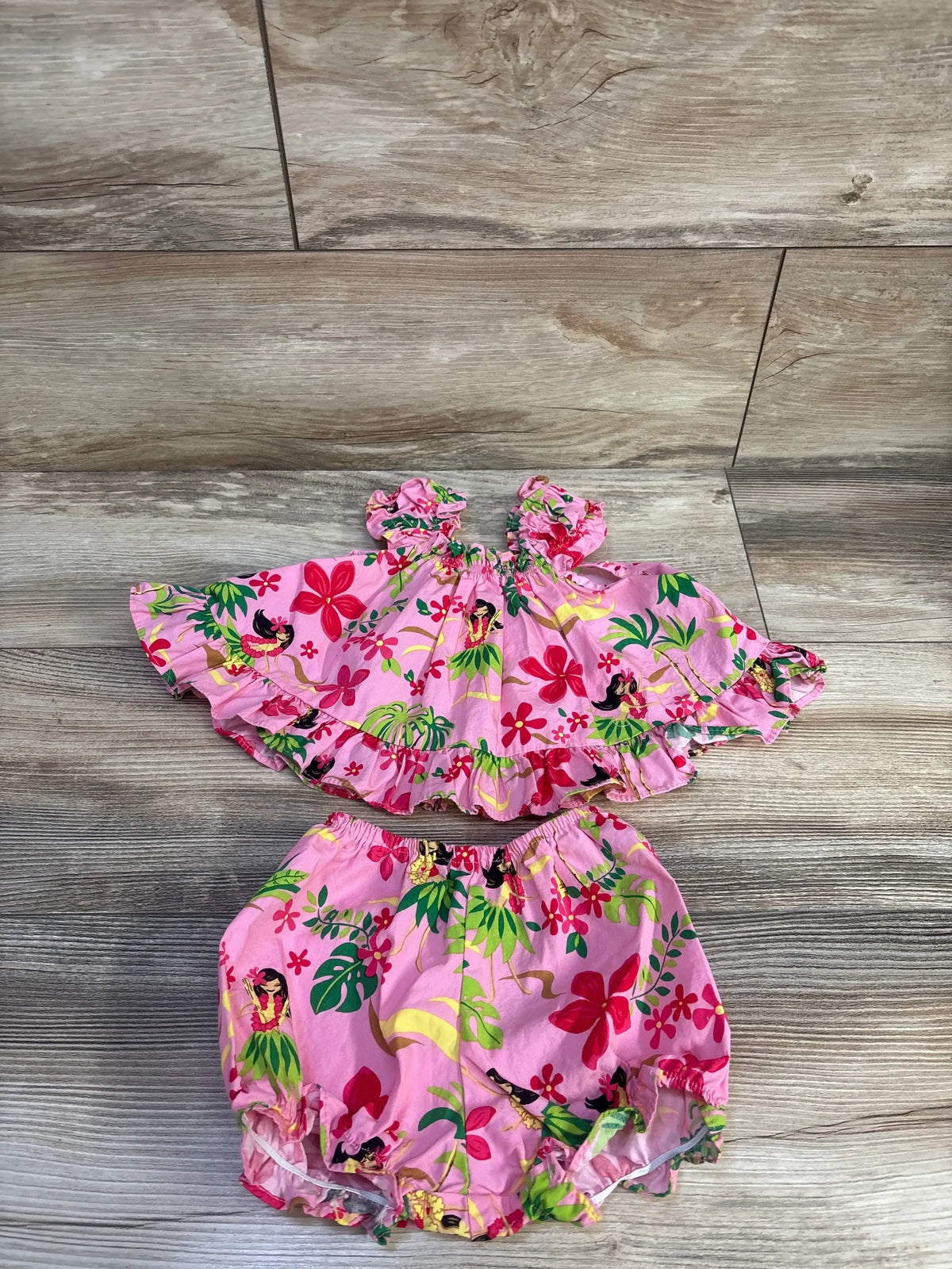 Flat lay of pink 2-piece cabana set: ruffled top and bloomers with tropical hula girl floral print on wood floor