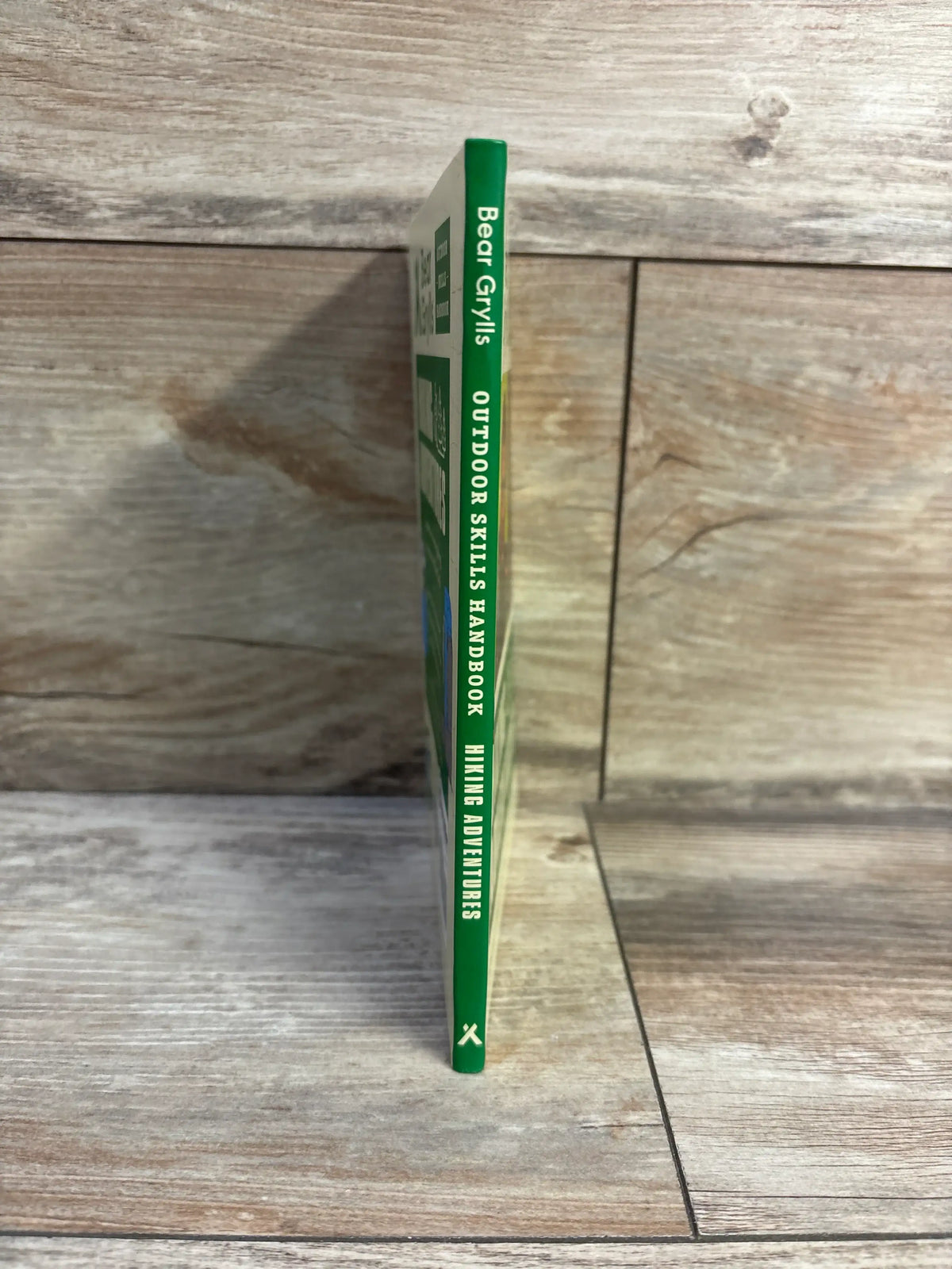 Spine view of Hiking Adventures Bear Grylls Outdoor Skills Handbook paperback showcasing its green spine with a wood-look background
