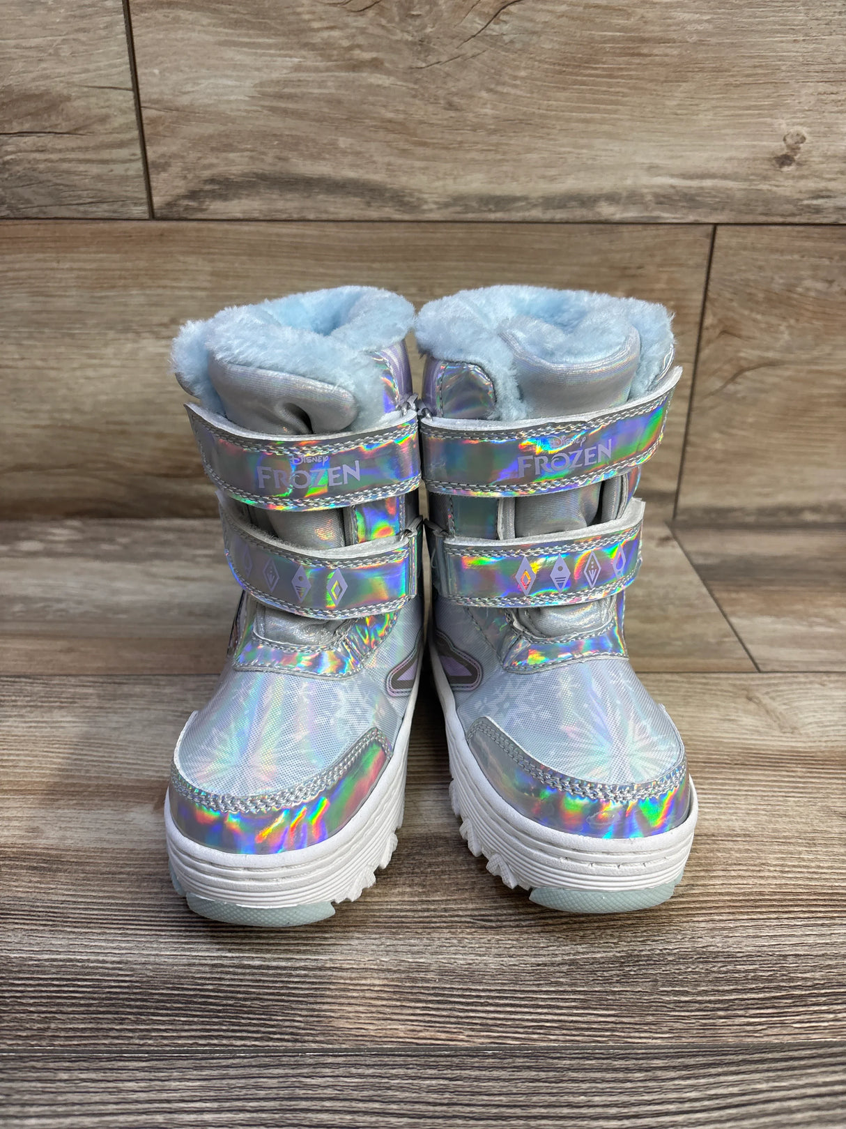 Front view of Disney Frozen toddler snow boots, silver holographic upper with three ‘FROZEN’ velcro straps and blue faux-fur lining on wood floor