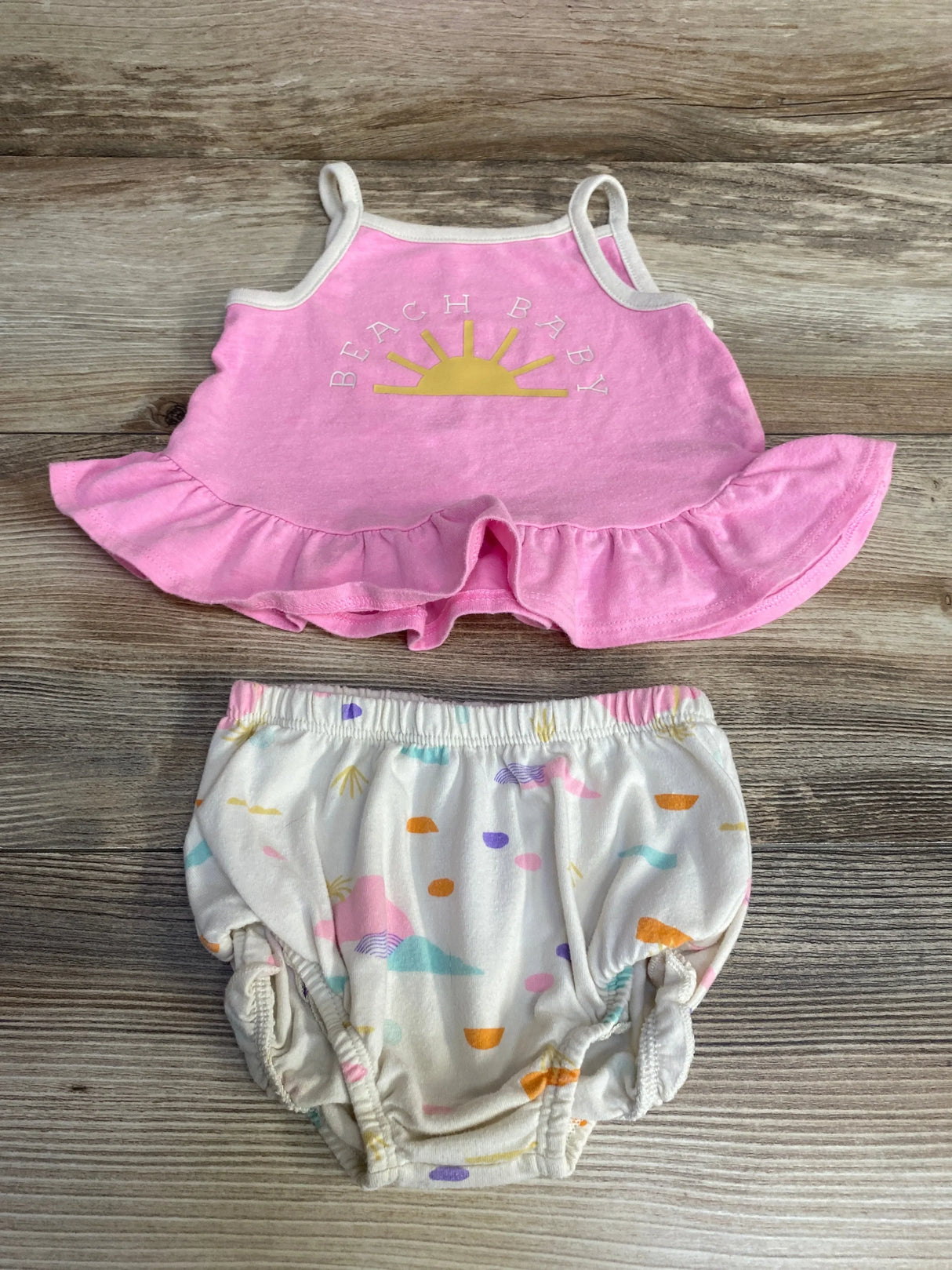 Top-down of Cat and Jack pink ruffle-hem baby tank with BEACH BABY sun graphic and white pastel bloomers on wood background