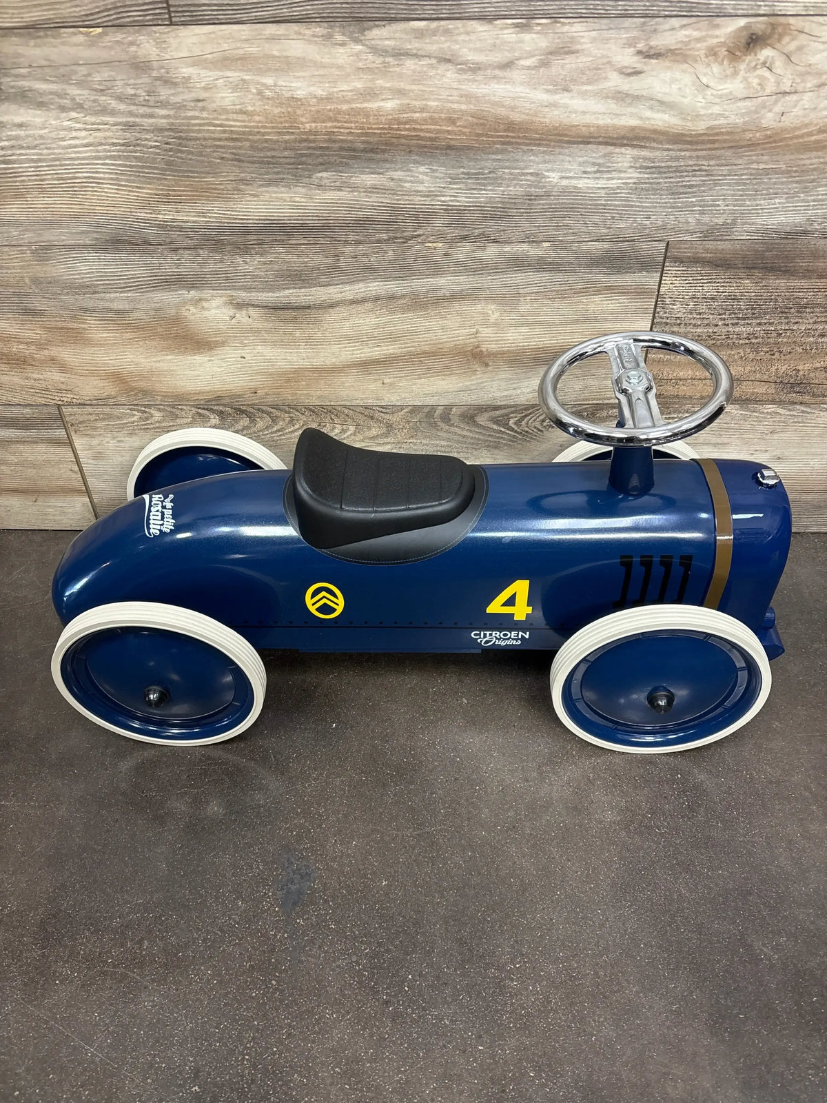 Side view of Baghera Speedster Rosalie Citroen ride-on in glossy blue showing black seat, white-trim wheels, yellow number 4 and chrome steering wheel on dark floor with wood-panel backdrop