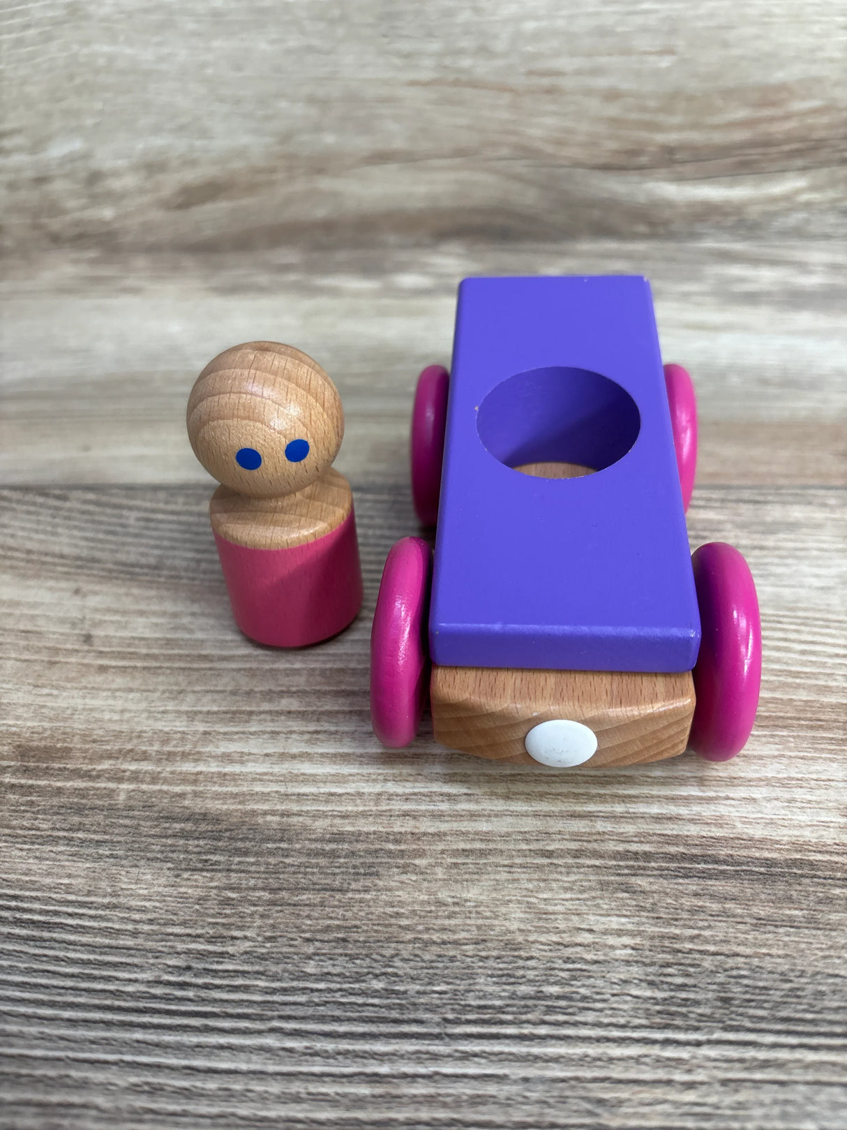 Wooden peg doll with pink body next to a purple and pink wooden toy car on wooden surface