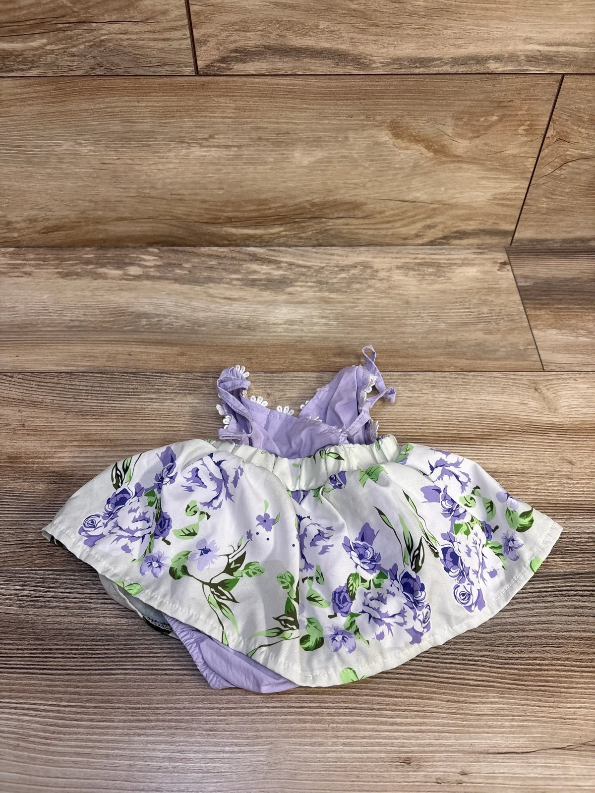 Top-down flat lay of purple floral baby bodysuit dress size 3-6 months showing ruffled shoulder straps, elastic waist, and attached skirt on wood floor