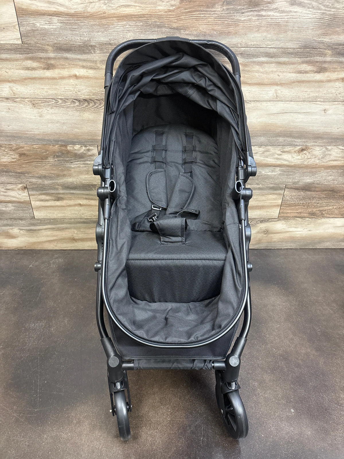 Front view of black Cochilder Lite 2.0 stroller showcasing padded seat and safety harness with wood-look flooring