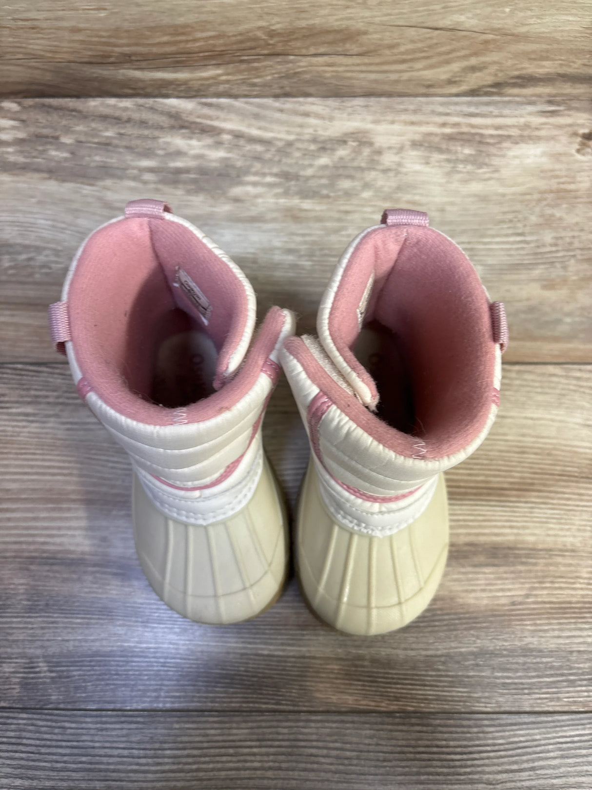 Top-down view of Oshkosh toddler splash winter boots showcasing ivory rubber shell, quilted ivory upper, pink interior lining and pull tabs on a wood floor