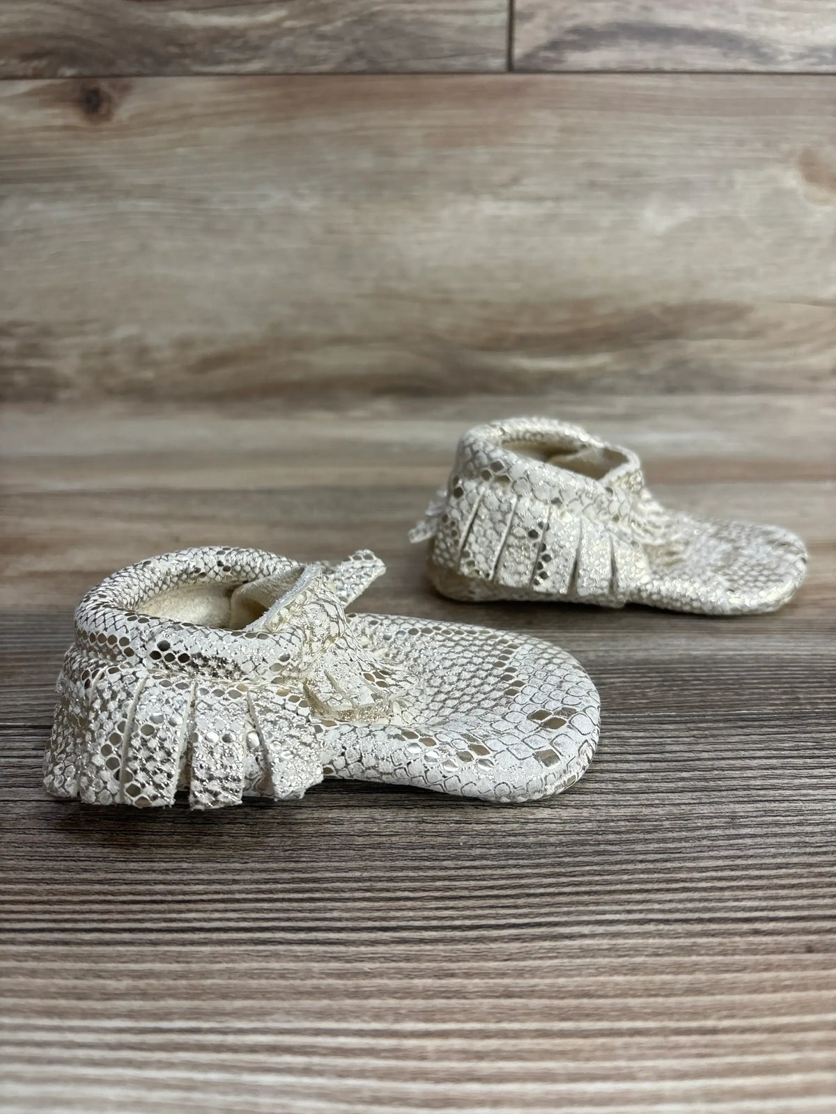 Pair of white snakeskin-patterned baby moccasin shoes with fringe on wooden surface
