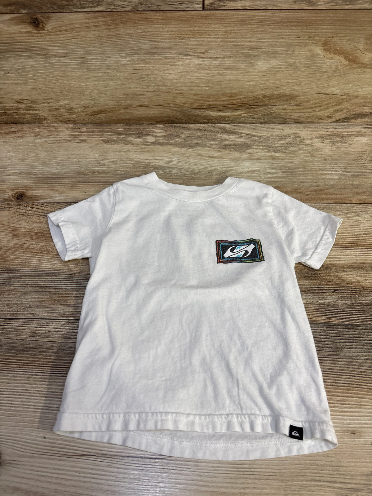 Flat lay of white Quiksilver toddler T-shirt with small embroidered chest logo and black hem tag on light wood background