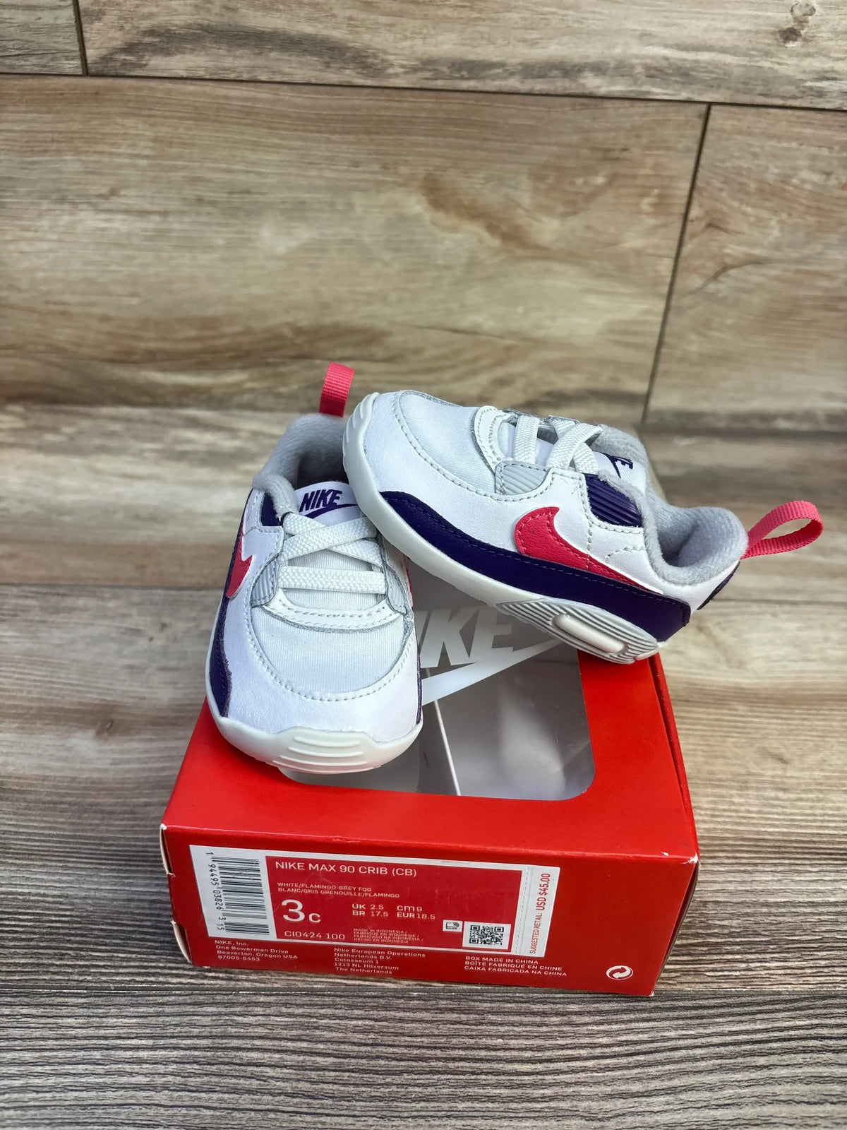 Three-quarter view of Nike Air Max 90 crib sneakers size 3C showing white uppers, pink Swoosh and purple accents on red Nike box