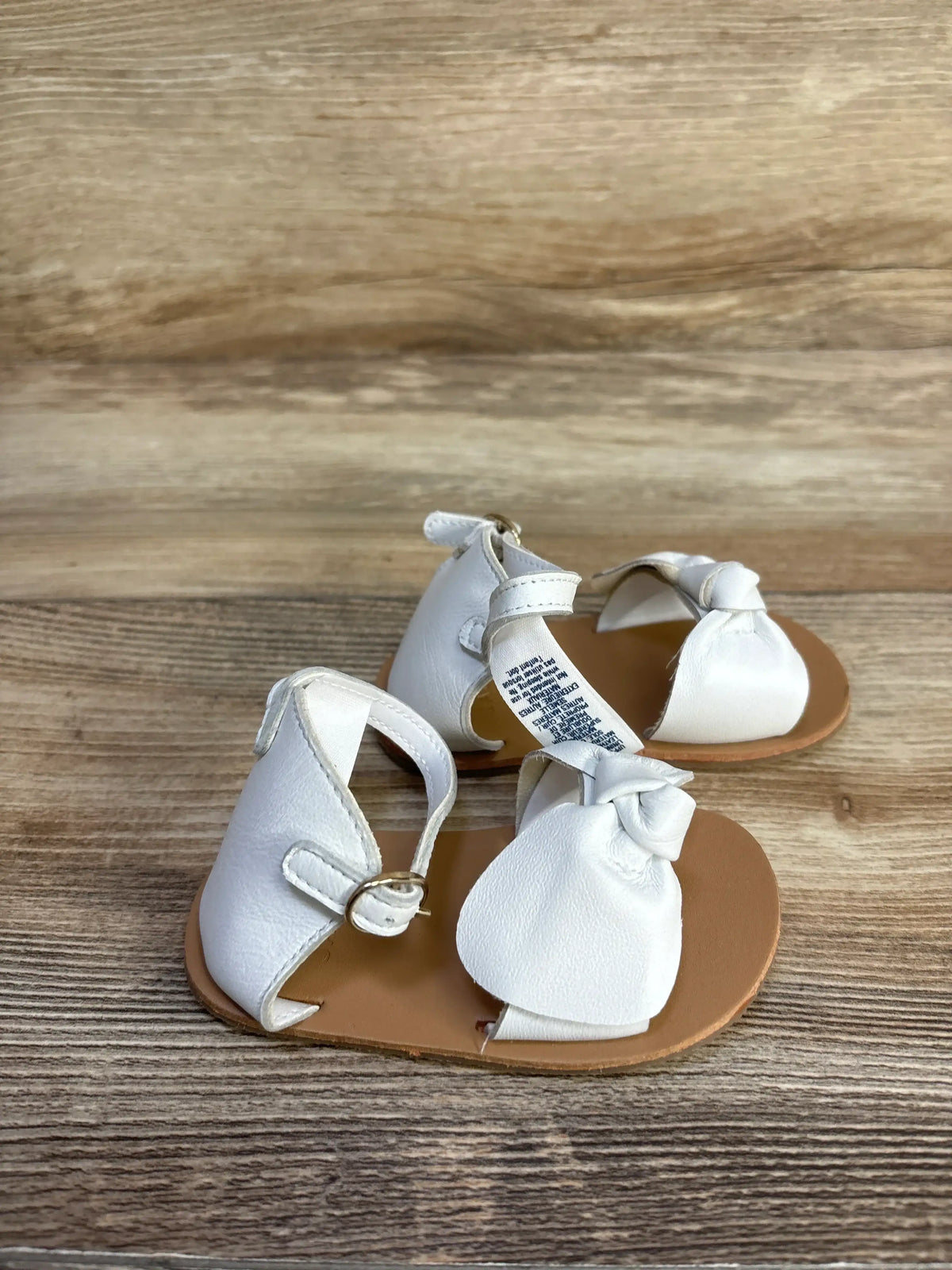 Angled view of white leather baby sandals showcasing knotted bow and ankle strap on wood background