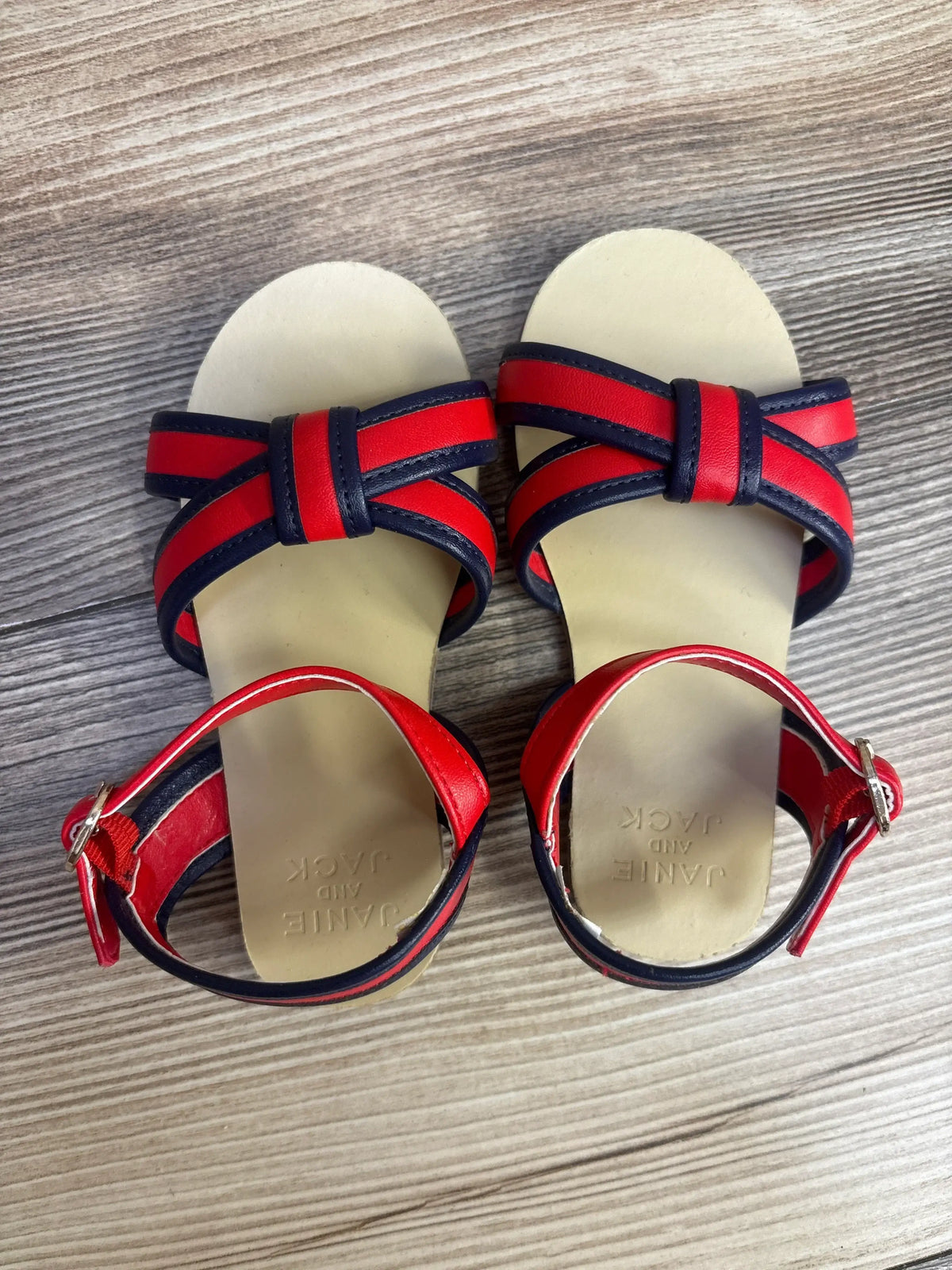 Top-down view of Janie and Jack toddler cross-strap sandals showing red and navy striped straps, ankle buckle, and cream footbed on wood floor