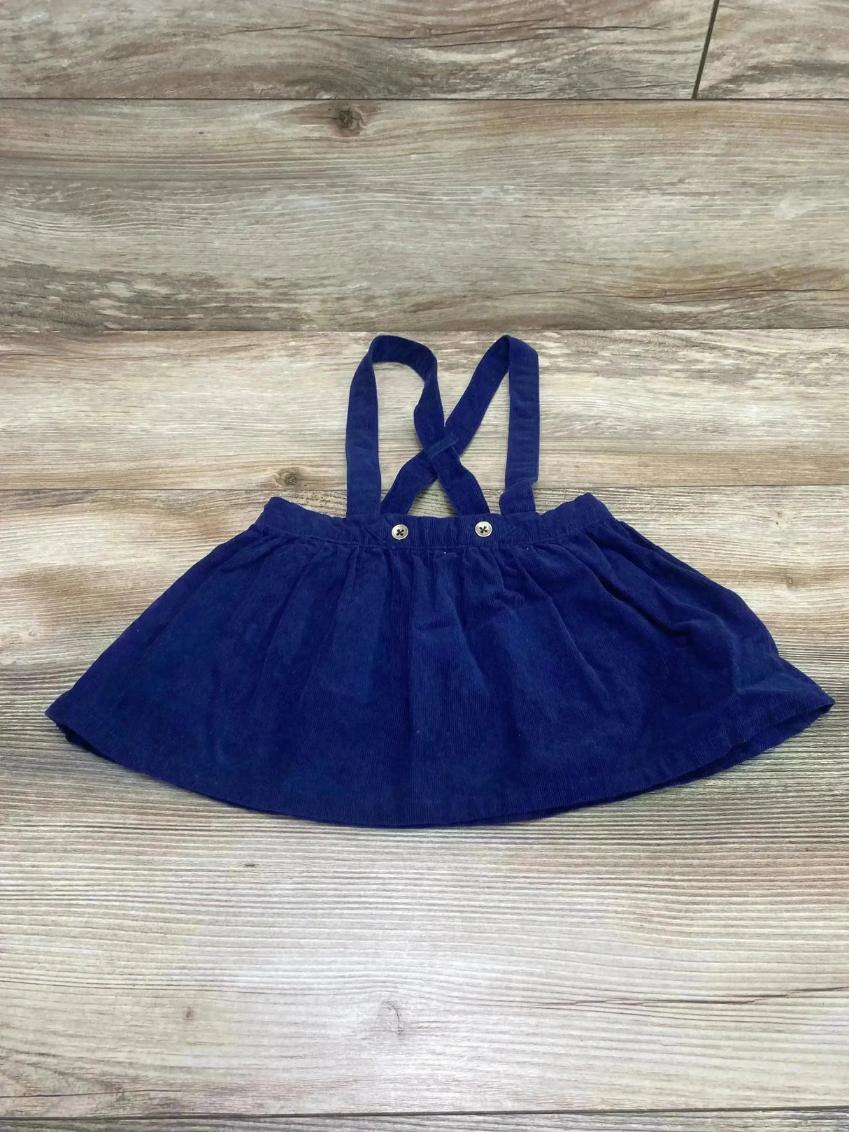 Flat lay of Cat and Jack Corduroy Suspender Skirt Navy sz 12m showcasing navy corduroy skirt with crossed straps on wood plank floor