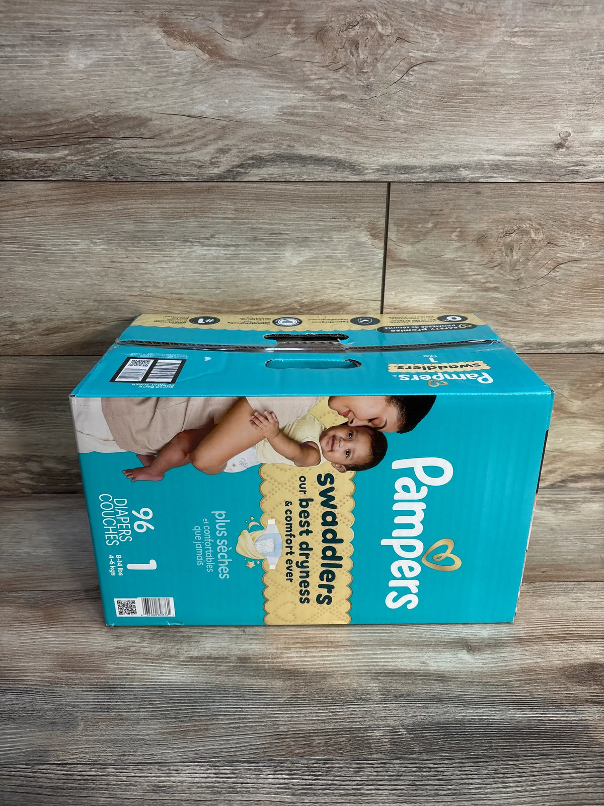 Front view of Pampers Swaddlers Size 1 diaper box showcasing 96 diapers and dryness and comfort messaging on a wood floor