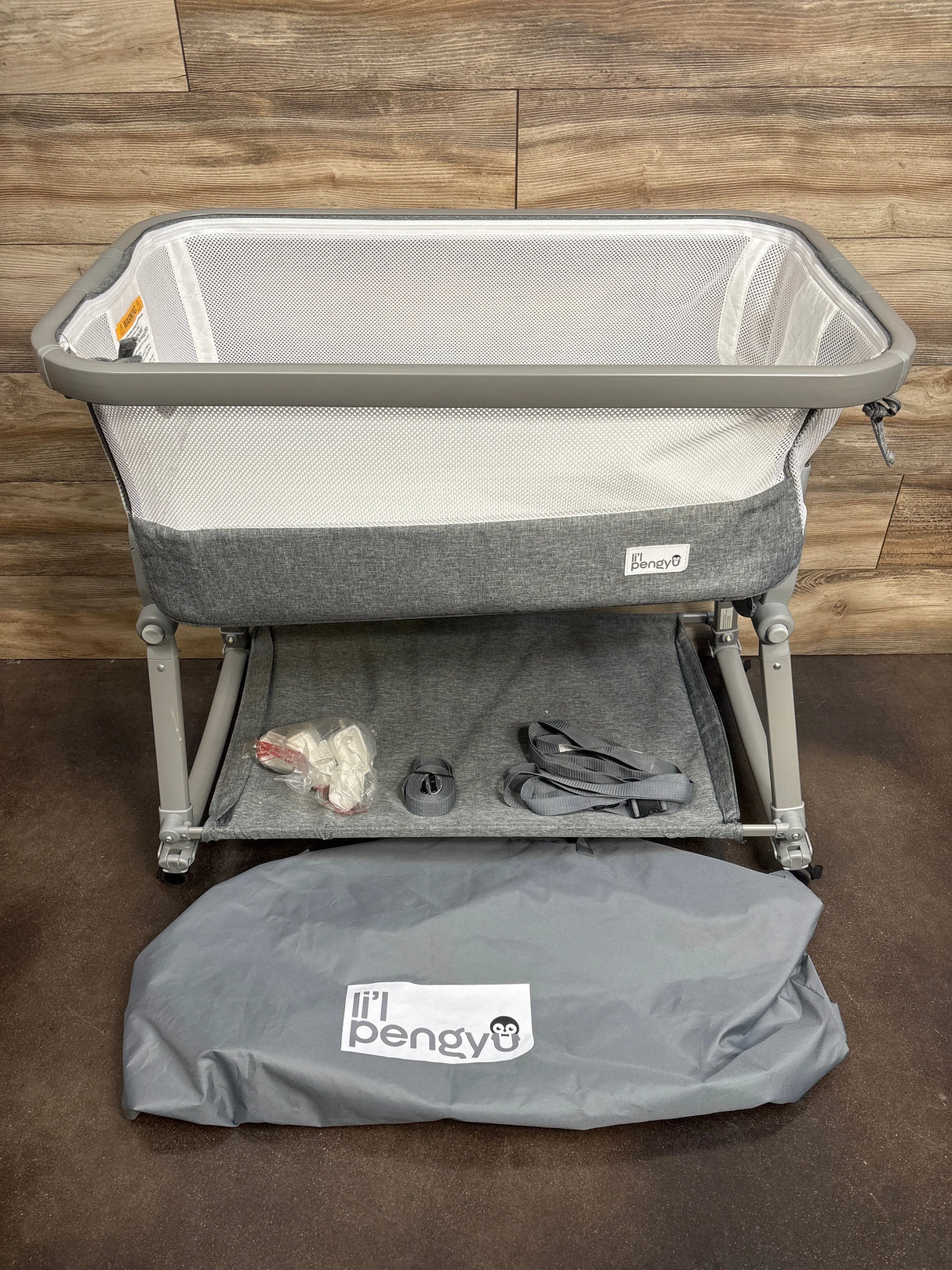 Gray mesh baby bassinet with accessories and 'li'l pengyu' storage bag on wooden floor
