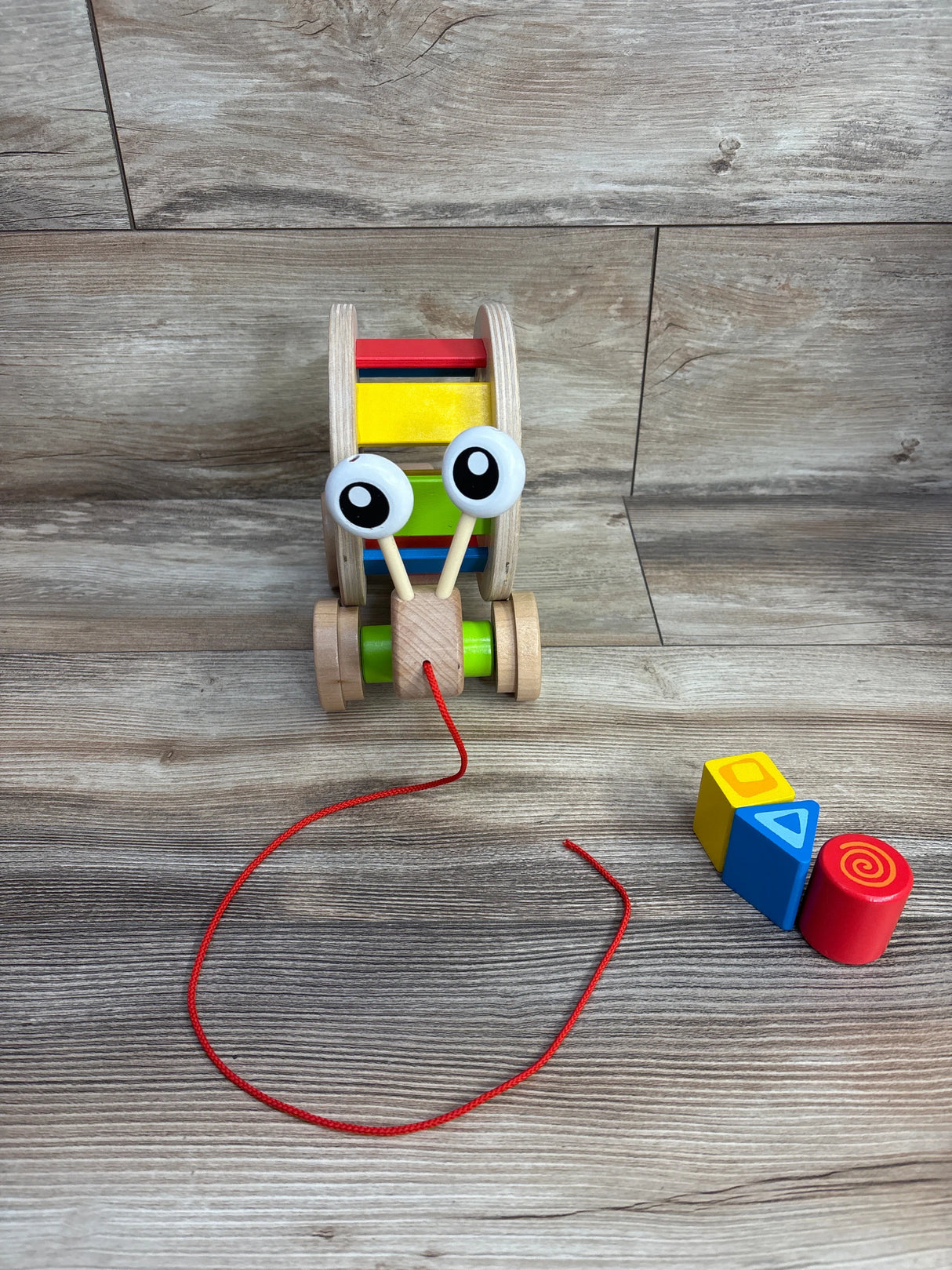 Wooden pull-along toy with large eyes and colorful rolling drum beside three colorful stacking blocks on wood surface