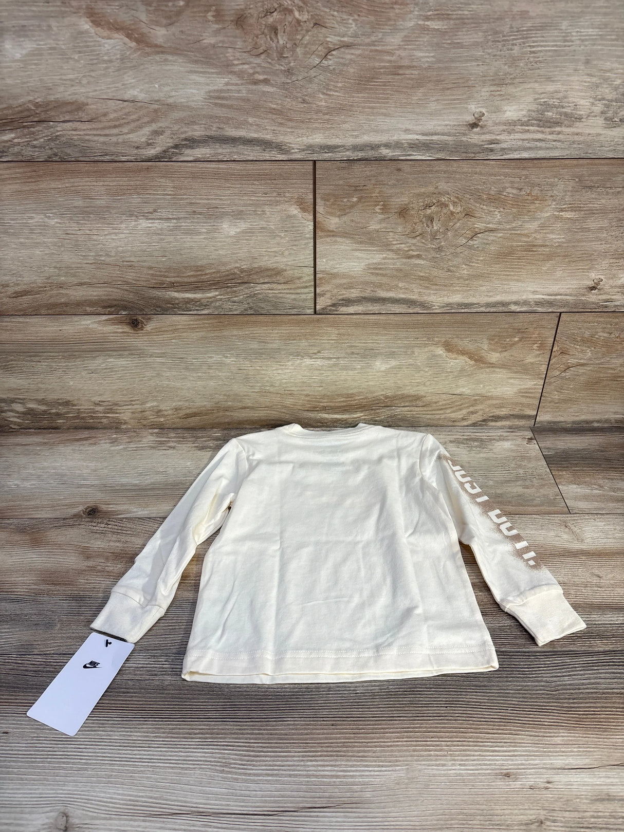 Back view of Nike 2T long-sleeve toddler shirt in coconut cream showing sleeve 'Just Do It' text on wood floor