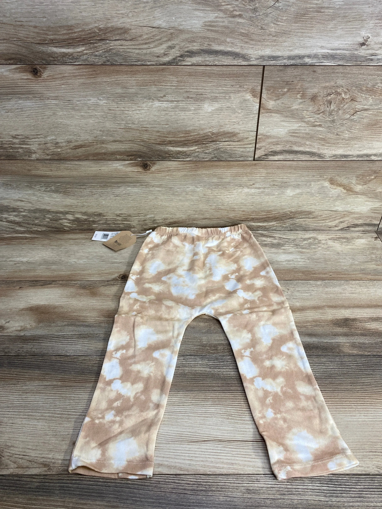 Flat lay of Grayson Mini joggers in tan and white tie-dye showcasing elastic waistband and brand hangtag on wood plank floor