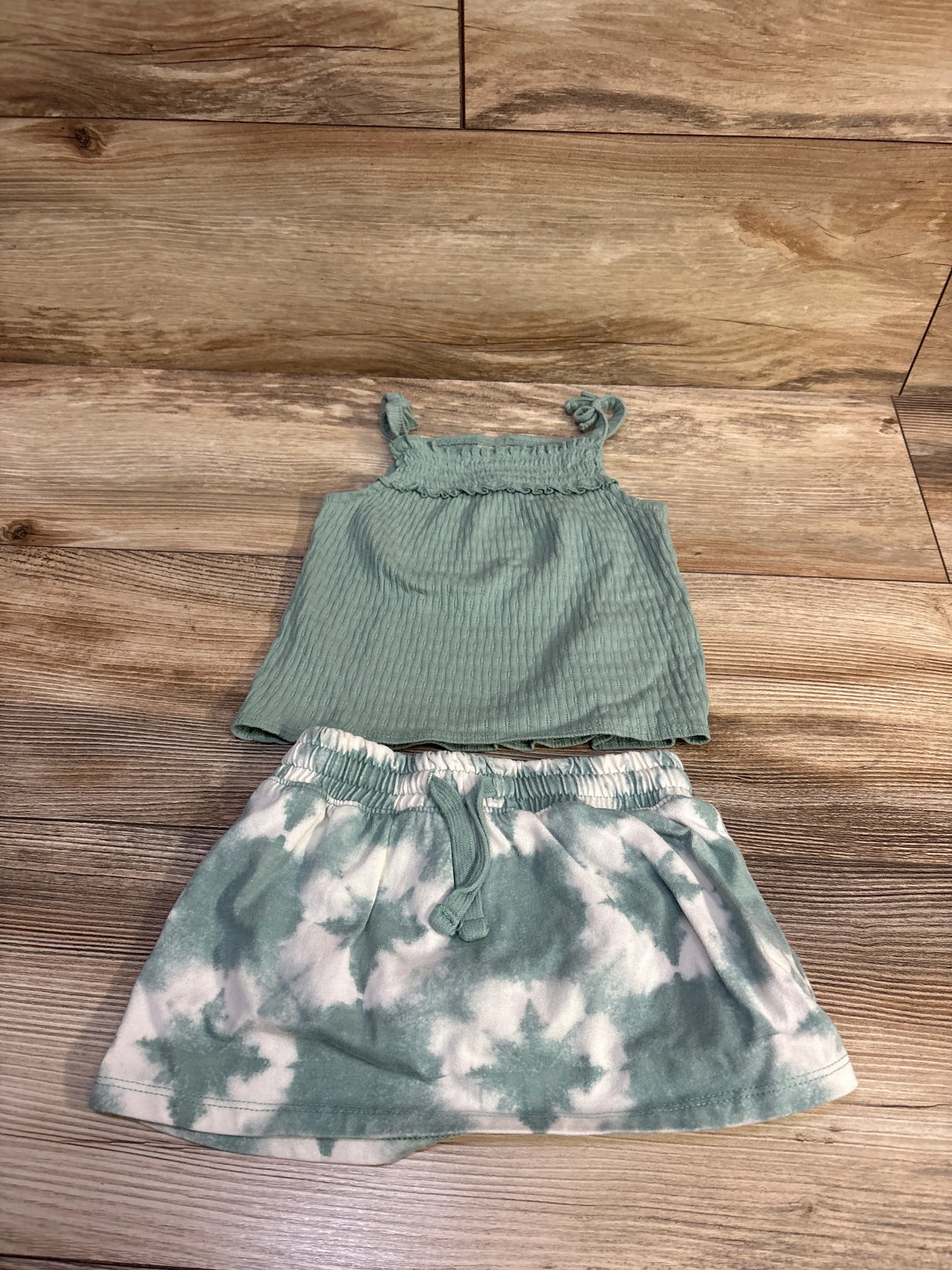 Flat lay of 2-piece green tank and tie-dye skort showing ribbed tank with knotted straps and drawstring waist on wood floor