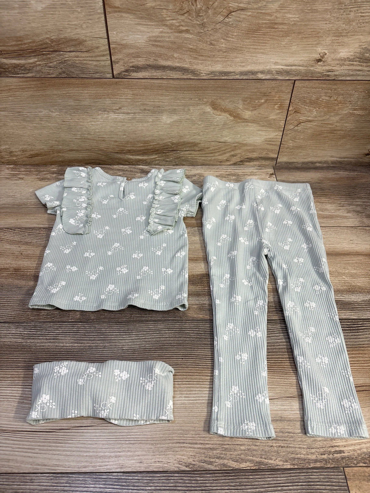 Flat lay of Calvin Klein 3-piece ribbed baby set with ruffle top, leggings and headband, white floral print on wood background