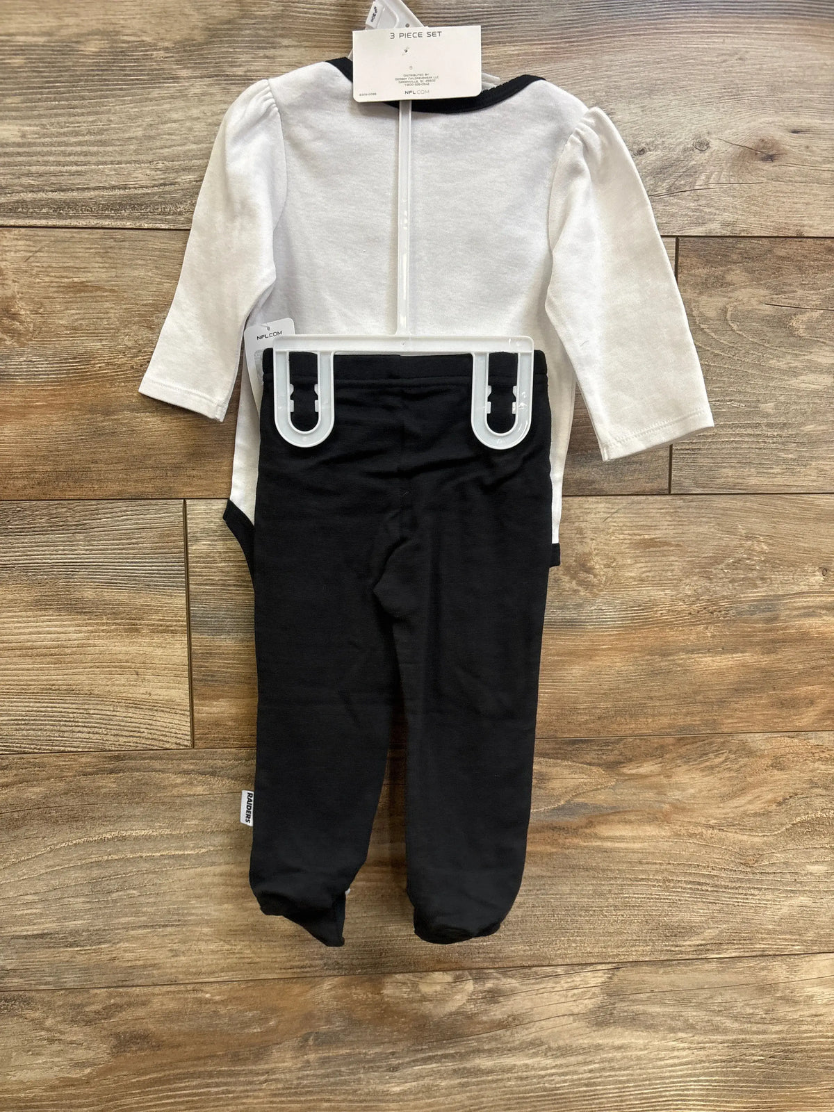 Back view of Las Vegas Raiders 3-piece baby bodysuit and footed pants set showcasing white long-sleeve bodysuit with black trim and black footed pants on wood floor