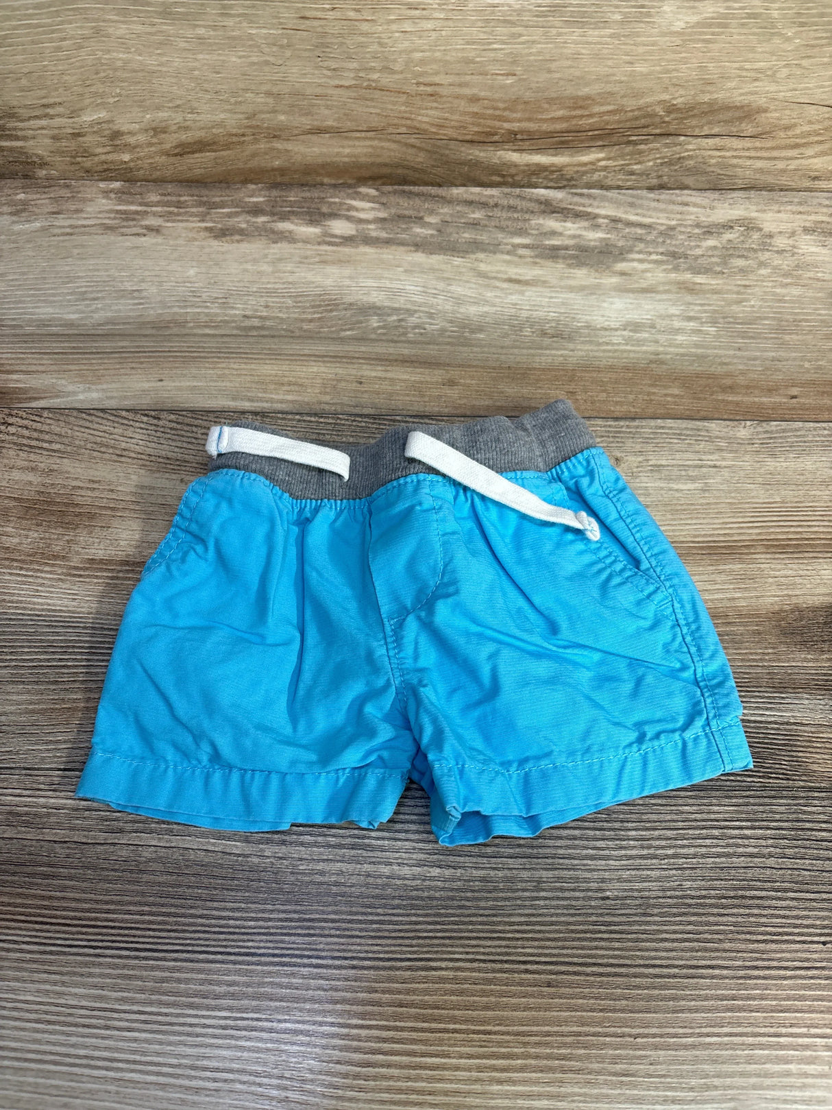 Flat lay of Carter's blue shorts 3-6M showing grey elastic waist with white drawstring and pockets on wood background