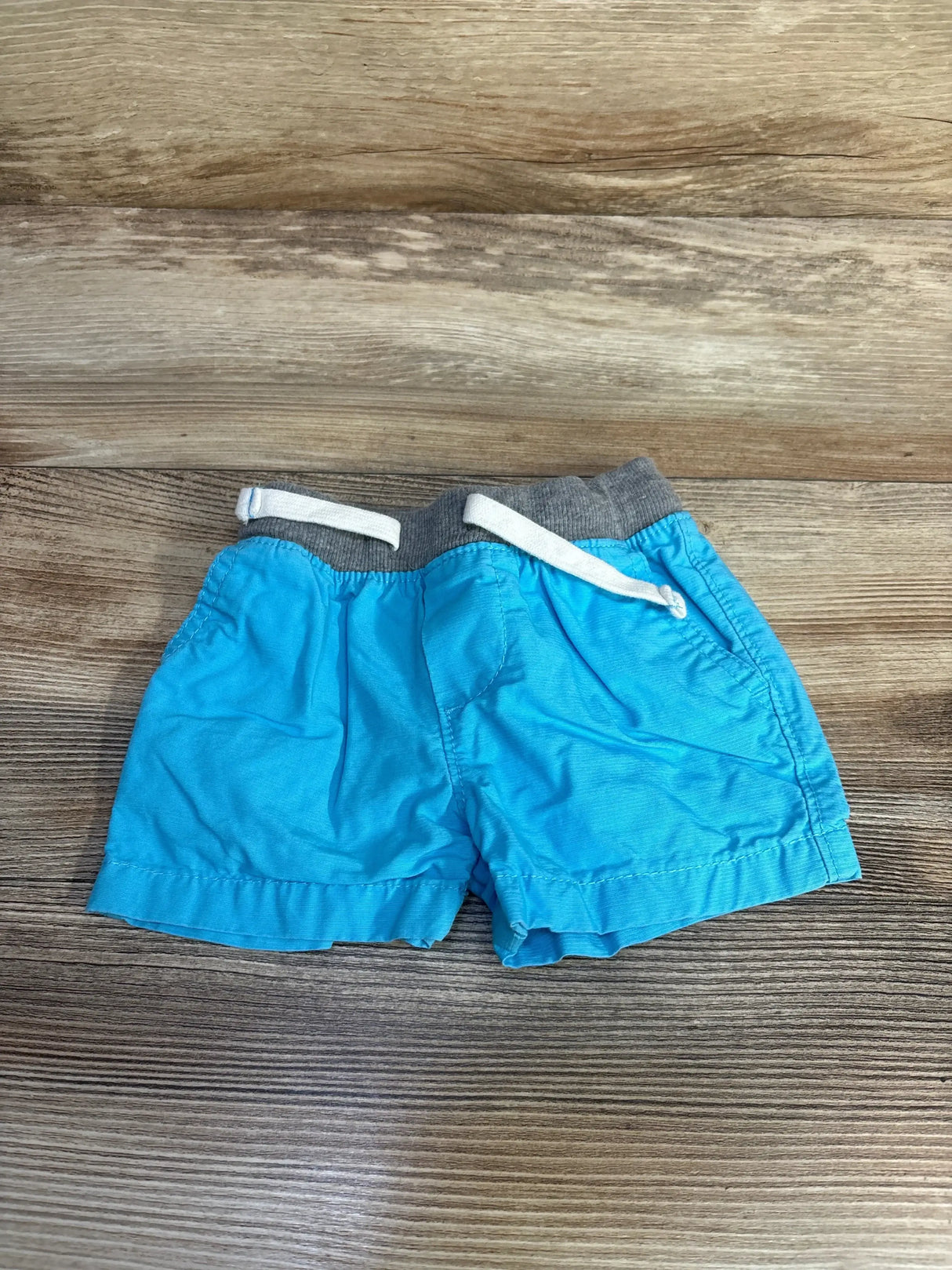 Flat lay of Carter's blue shorts 3-6M showing grey elastic waist with white drawstring and pockets on wood background
