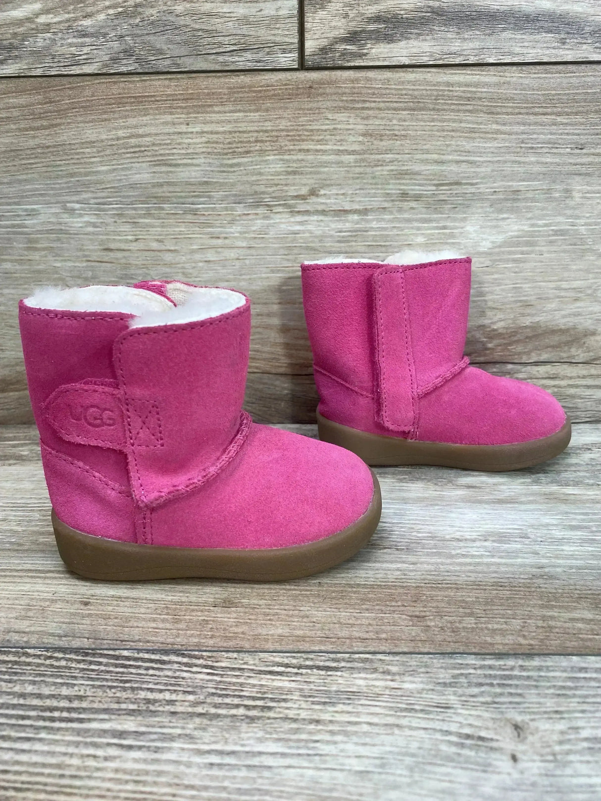 Side view of UGG Keelan toddler pink suede boots showcasing white shearling lining and brown rubber sole on wood plank background