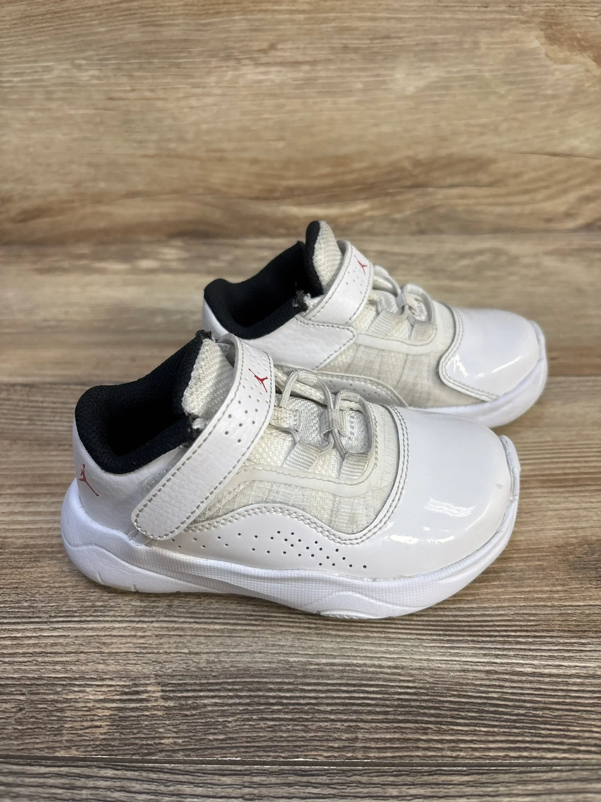 Side view of Air Jordan 11 CMFT Low GS ‘White Varsity Red’ toddler sneakers showcasing white patent-leather toe, Velcro strap with red Jumpman logo and black lining on wood floor