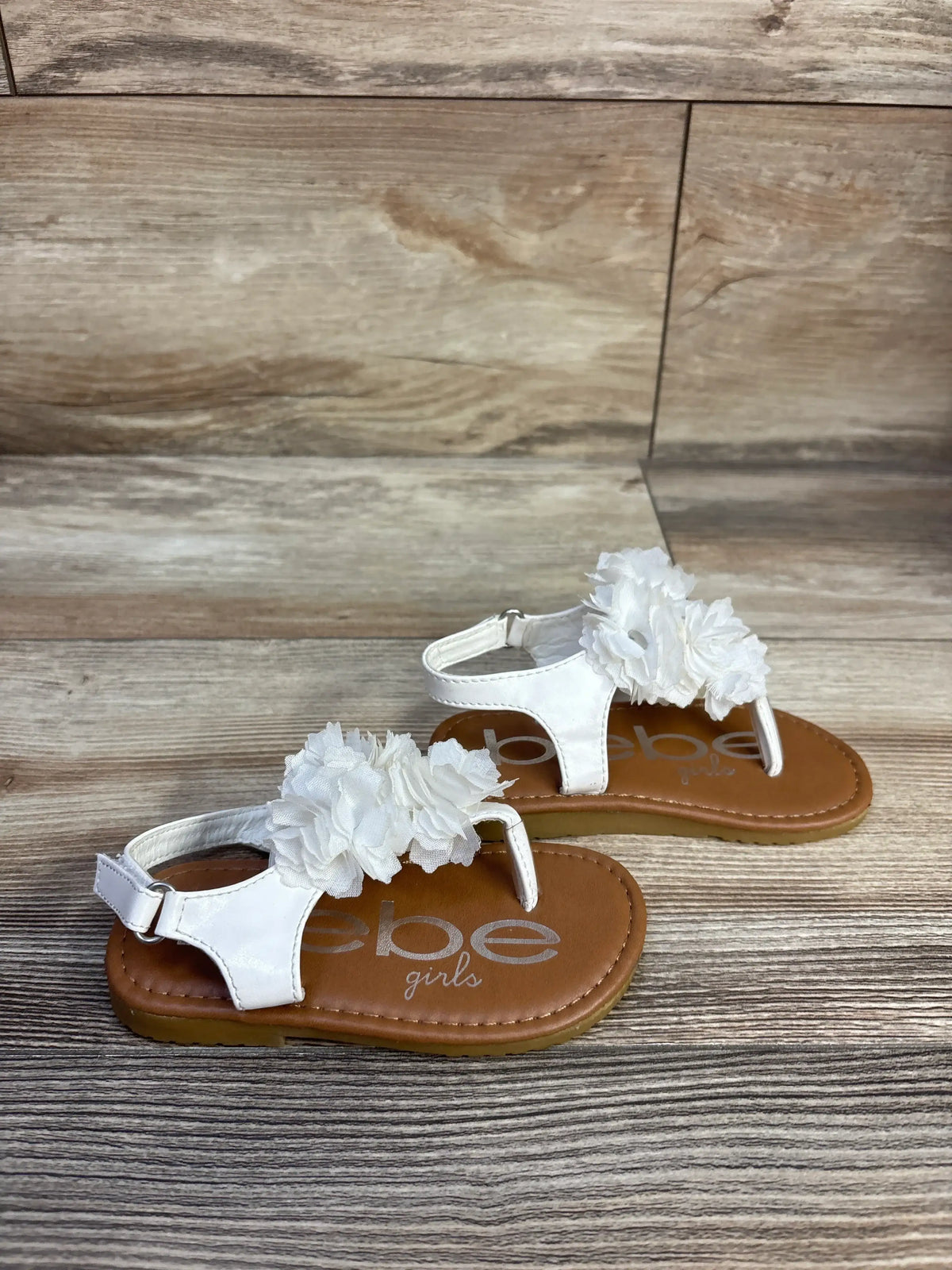 Angled view of Bebe white chiffon flower sandals in size 7C showcasing flower accents and buckle straps on a wood-look surface
