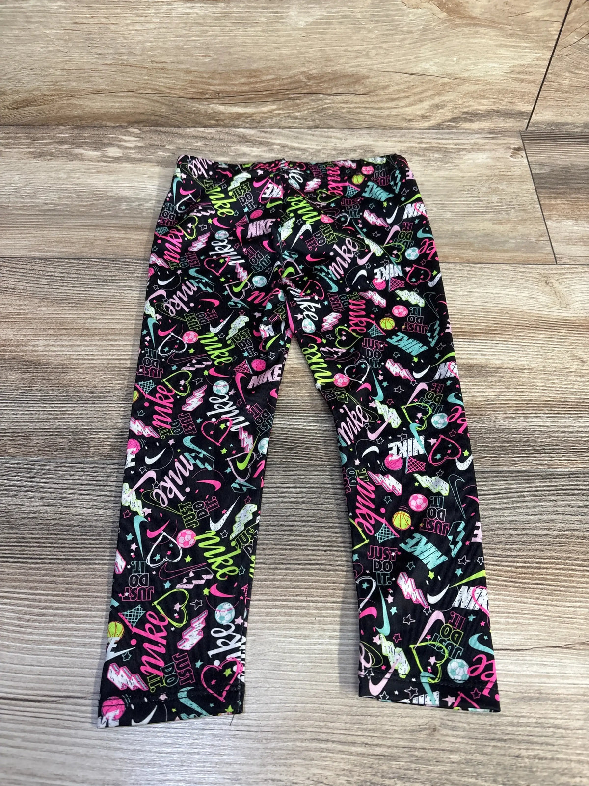 Flat lay of Nike toddler black leggings showcasing neon multicolor Nike logo and sports-themed allover print, size 24 months, on a wood floor