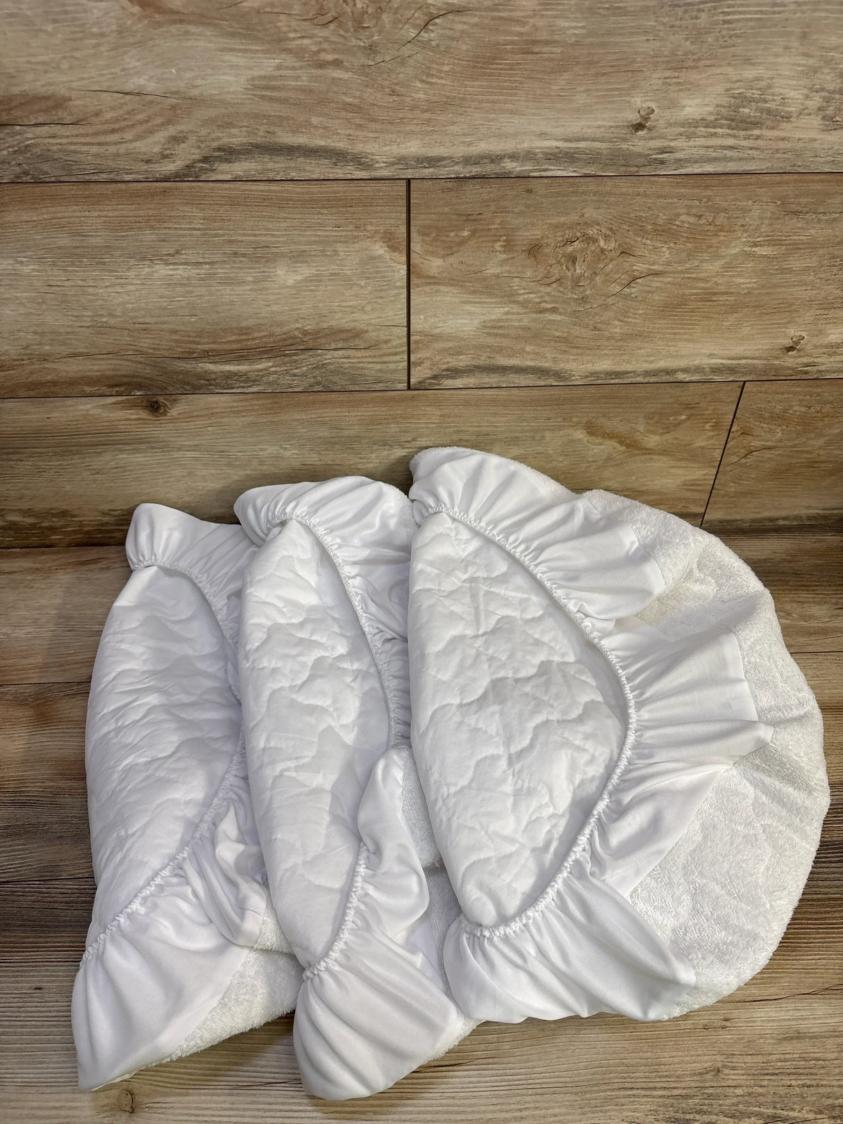 Top-down view of three white waterproof fitted bassinet sheets showing quilted surface and elastic fitted edges on wooden floor