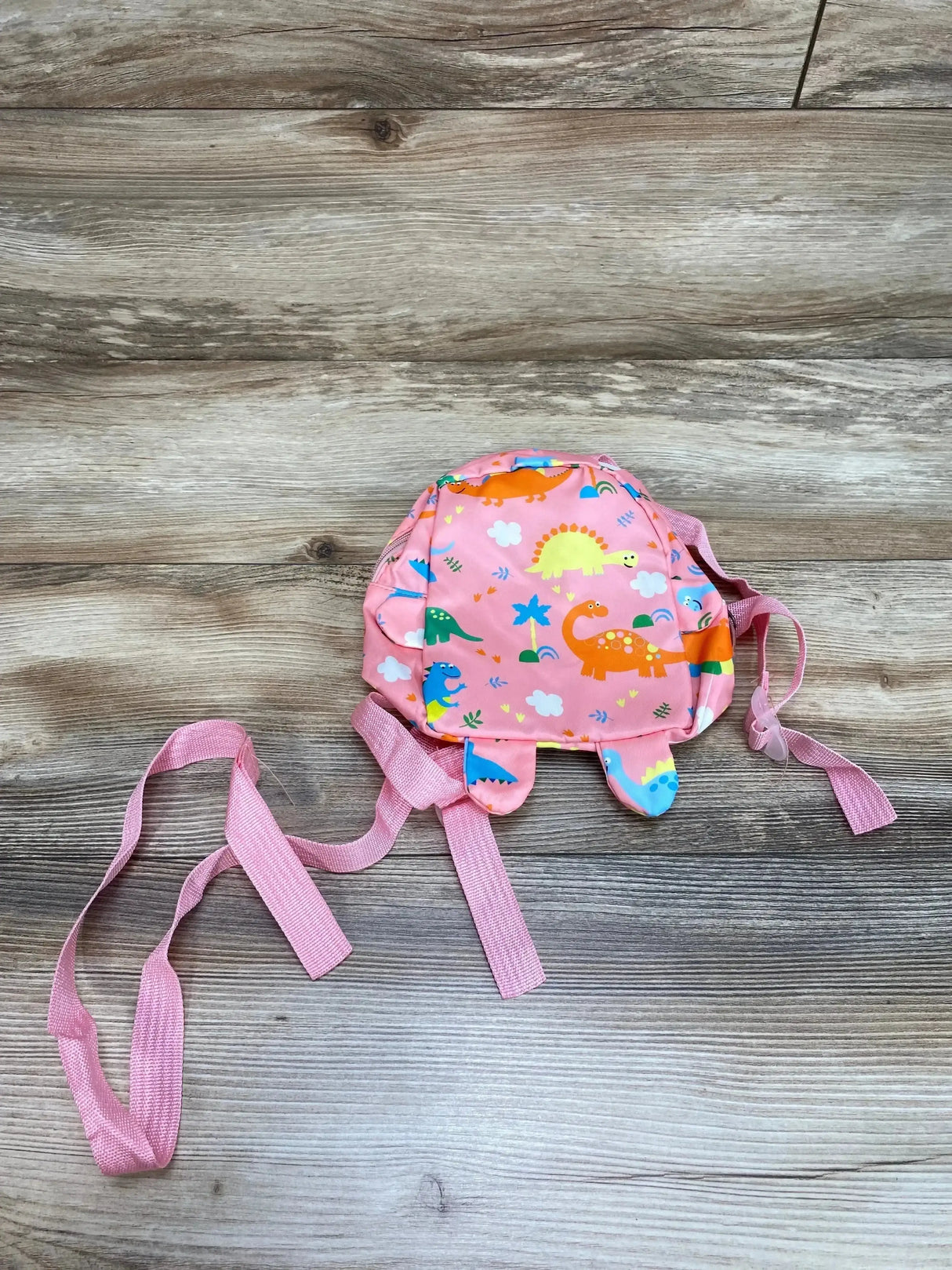 Top view of pink dinosaur safety harness backpack showcasing attached safety leash, adjustable straps, and dinosaur print on wood surface