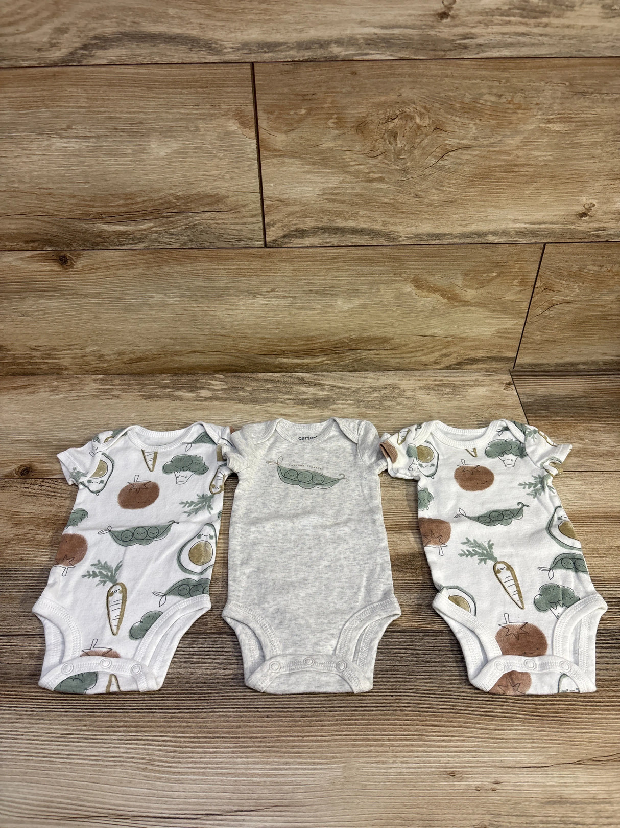 Top-down view of a 3-pack of newborn bodysuits showcasing vegetable prints and snap closures on a wood plank background