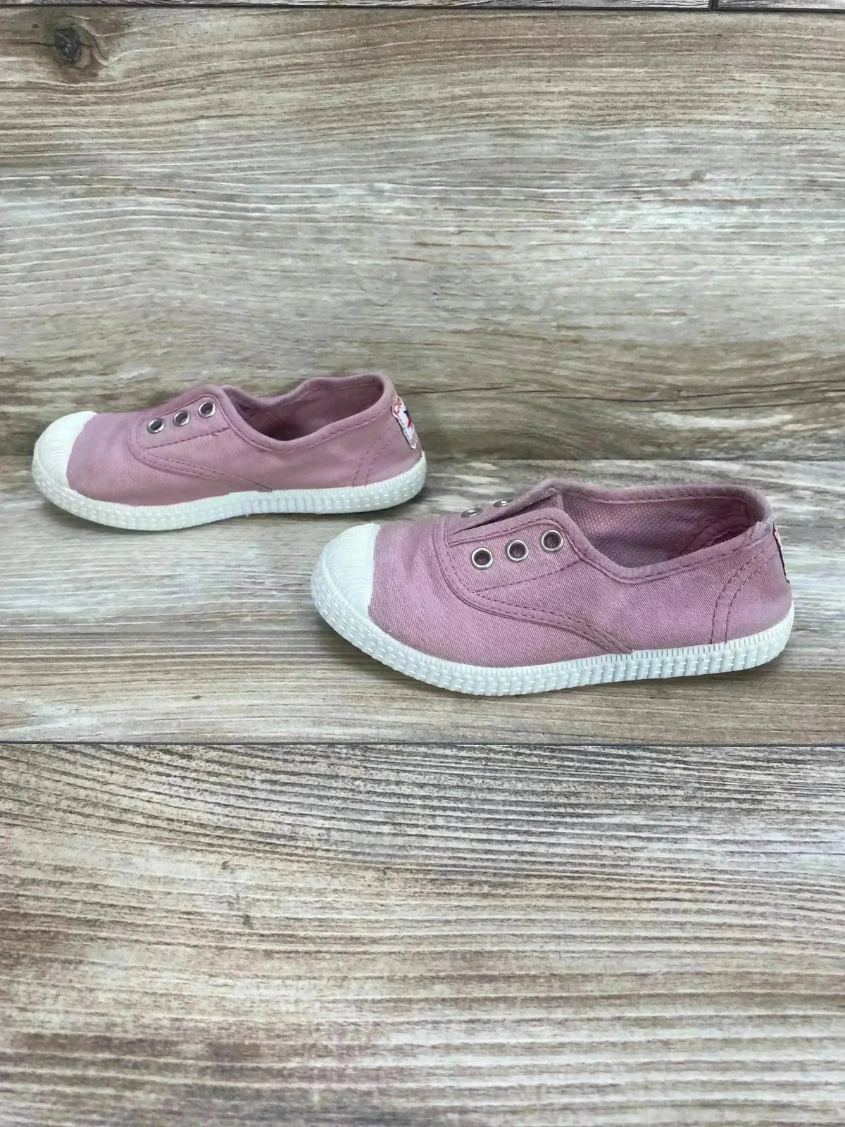 Side view of pink Cienta distressed canvas slip-on sneakers showcasing elastic openings and white soles on wood grain