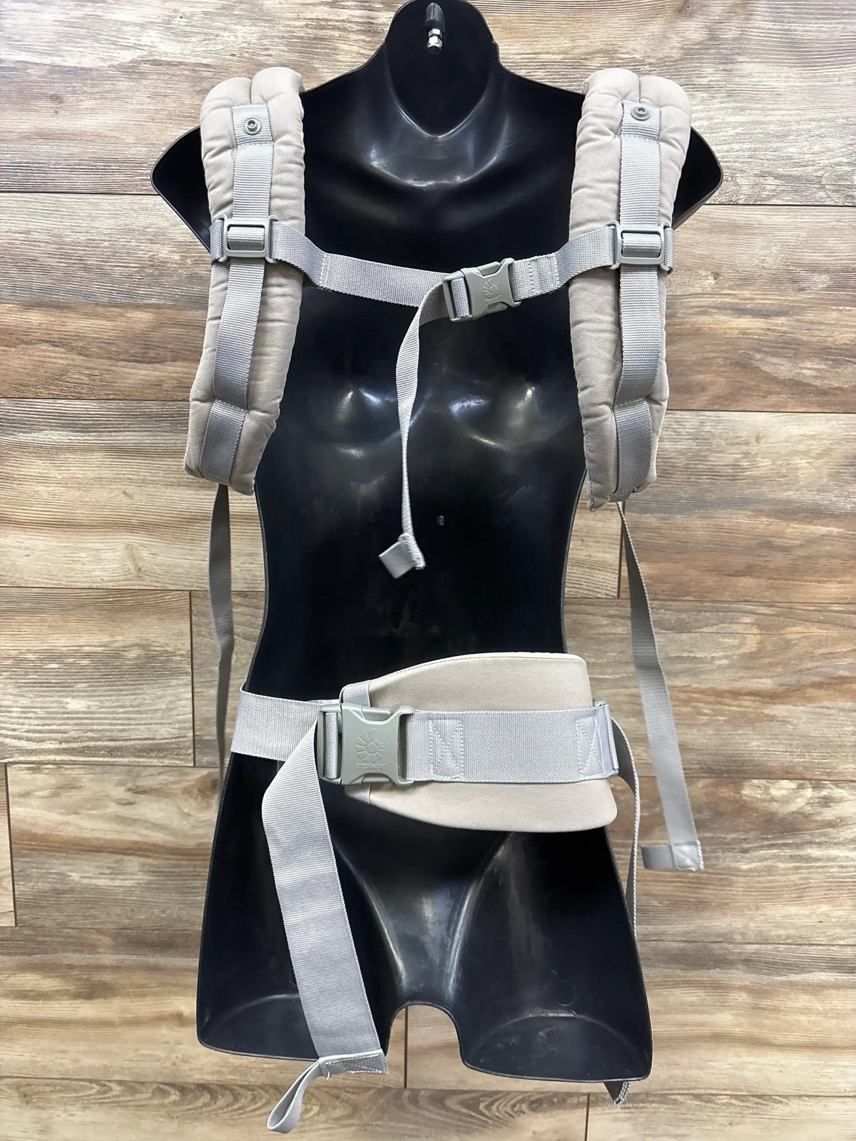 Back view of Ergobaby Omni 360 baby carrier in beige with padded straps and waist belt on black mannequin against wood backdrop