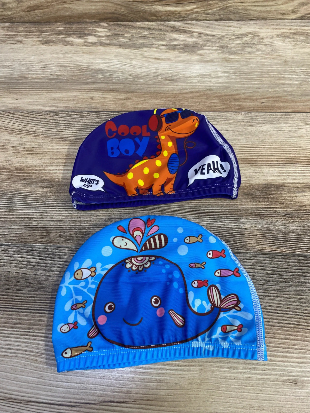 Top-down of two kids’ swim caps on wood: purple with orange dinosaur and ‘Cool Boy’ text; blue with smiling whale